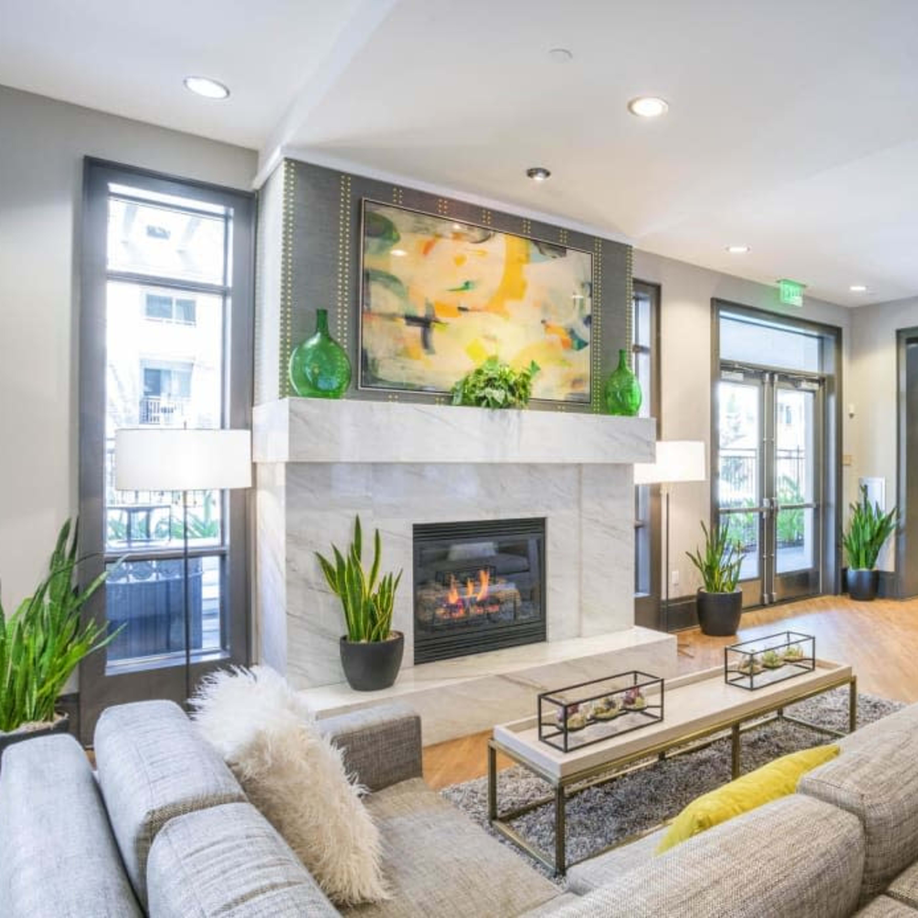 A modern living room features a fireplace, a large gray sectional sofa, and a vibrant painting above the mantel, complemented by plants and a kitchen area in the background.