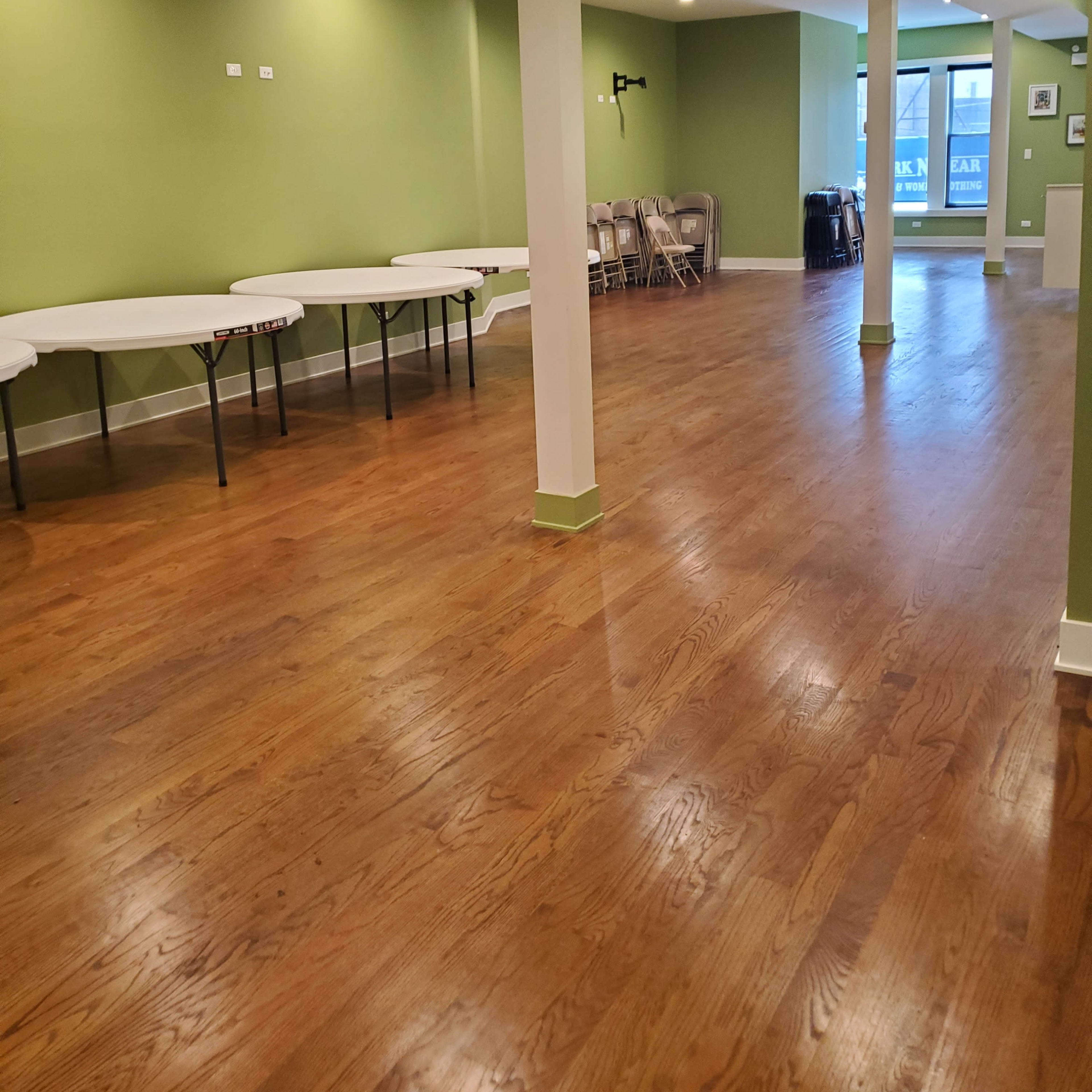 A spacious, green-walled room with wooden flooring, featuring several tables along one side and chairs stacked in the background.