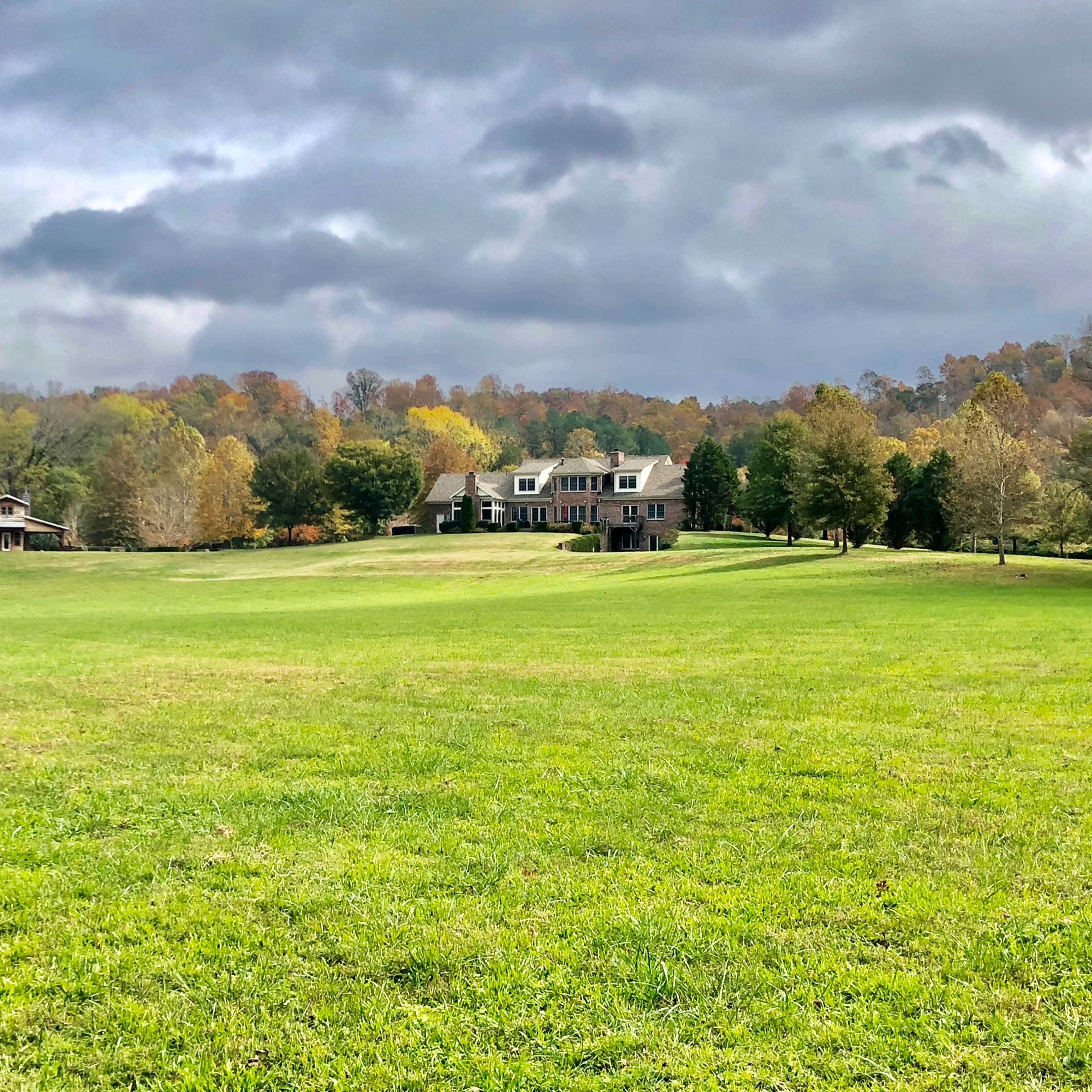 A large, two-story house is situated on a grassy field, surrounded by trees with changing foliage under a cloudy sky.