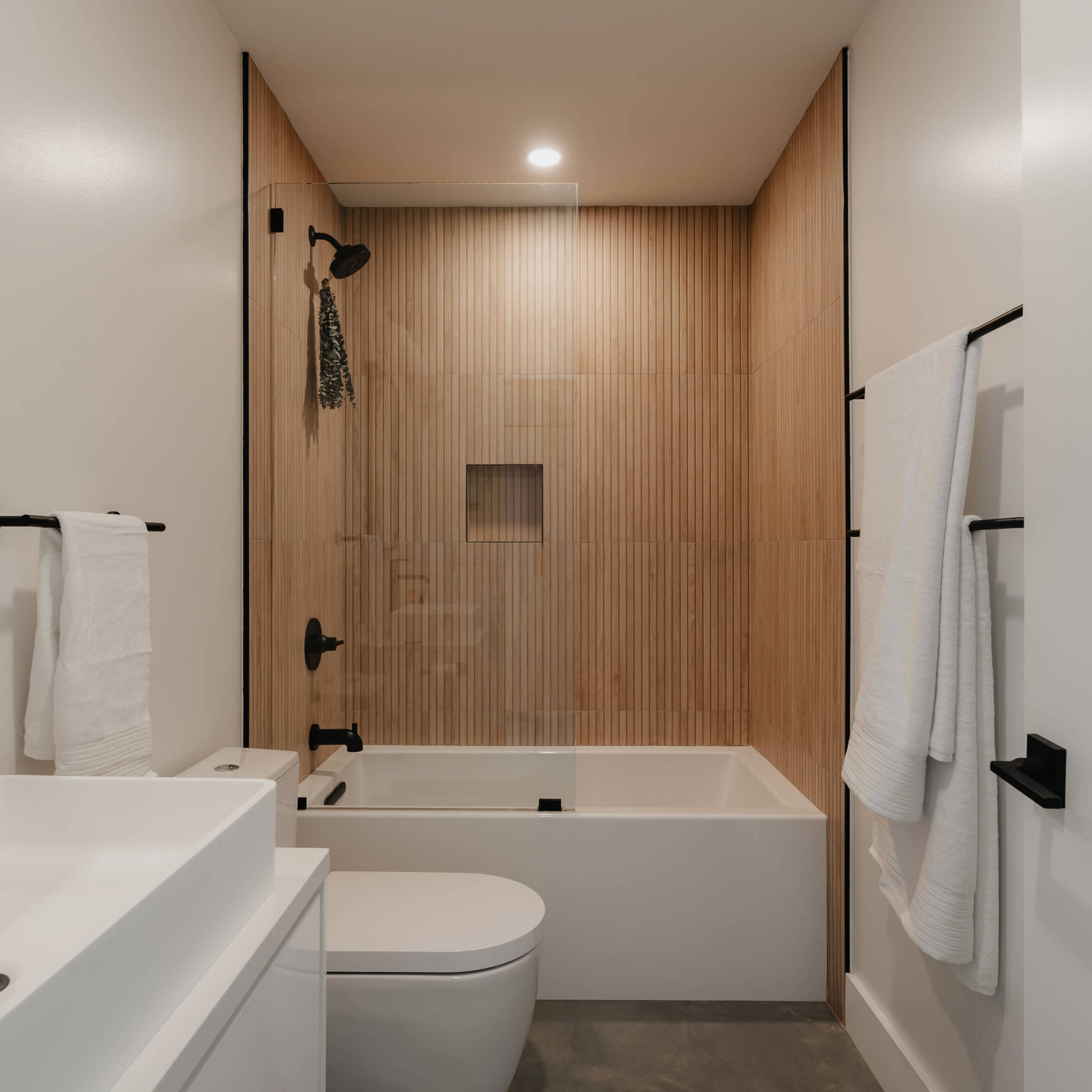 A modern bathroom featuring a bathtub with a shower, a minimalist vanity with a sink, and wooden paneling on the wall.