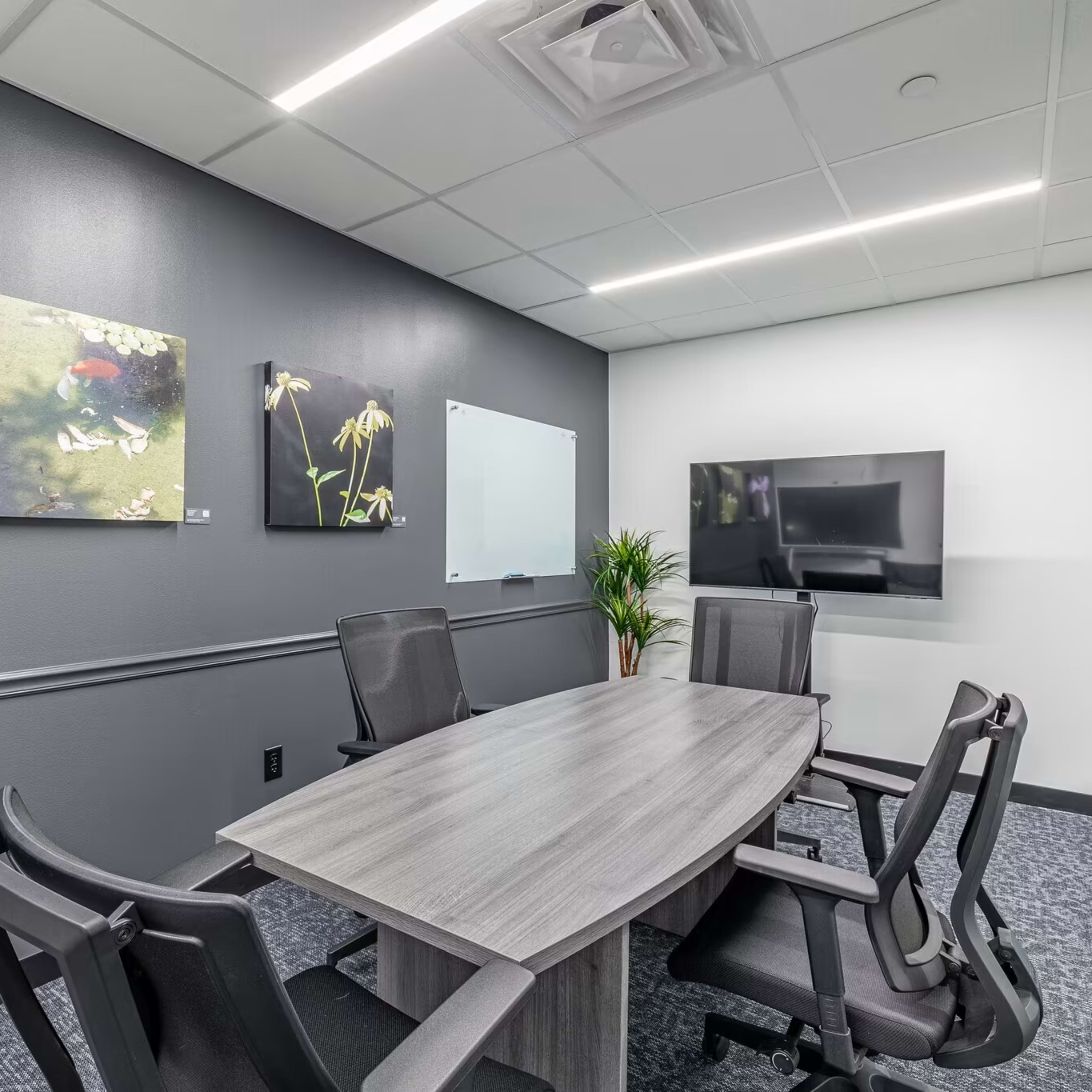 A modern meeting room features a large rectangular table surrounded by black chairs, framed artwork on the walls, a flat-screen TV, and a whiteboard.