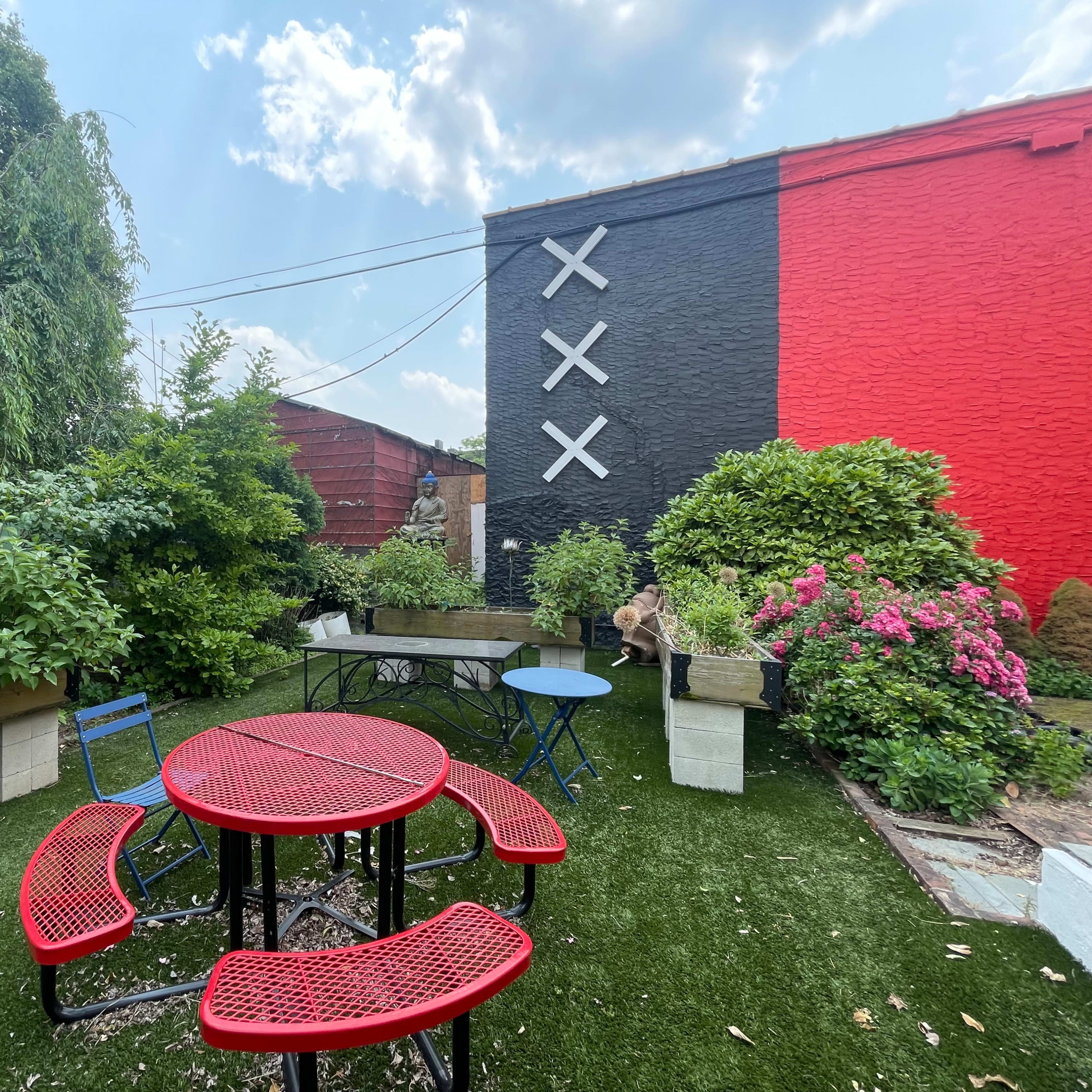 A landscaped outdoor area with red picnic tables, green plants, and a mural featuring black and red walls adorned with white cross symbols.