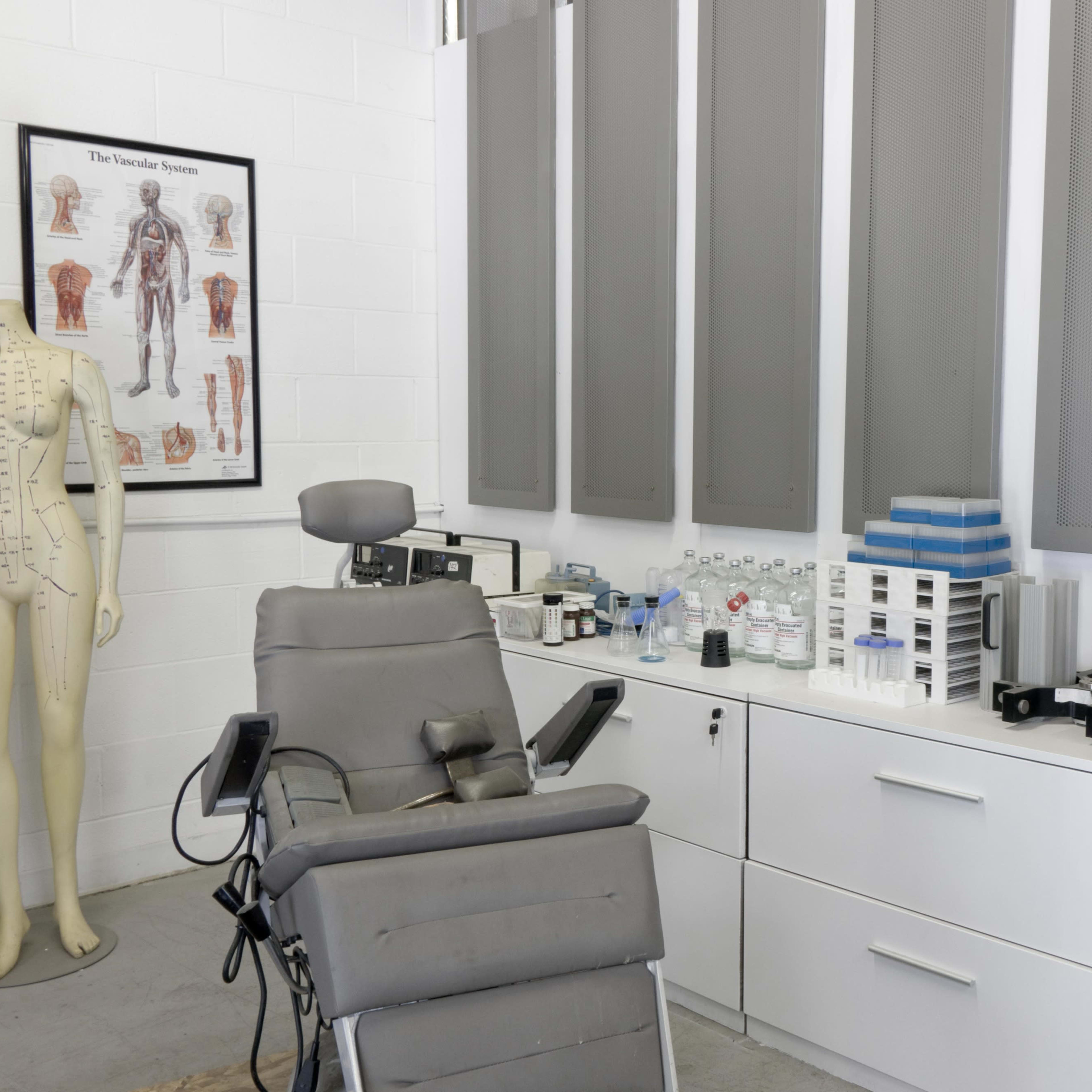 A medical examination room featuring a reclining chair, a mannequin, various medical supplies on shelves, and a wall chart of the human anatomy.