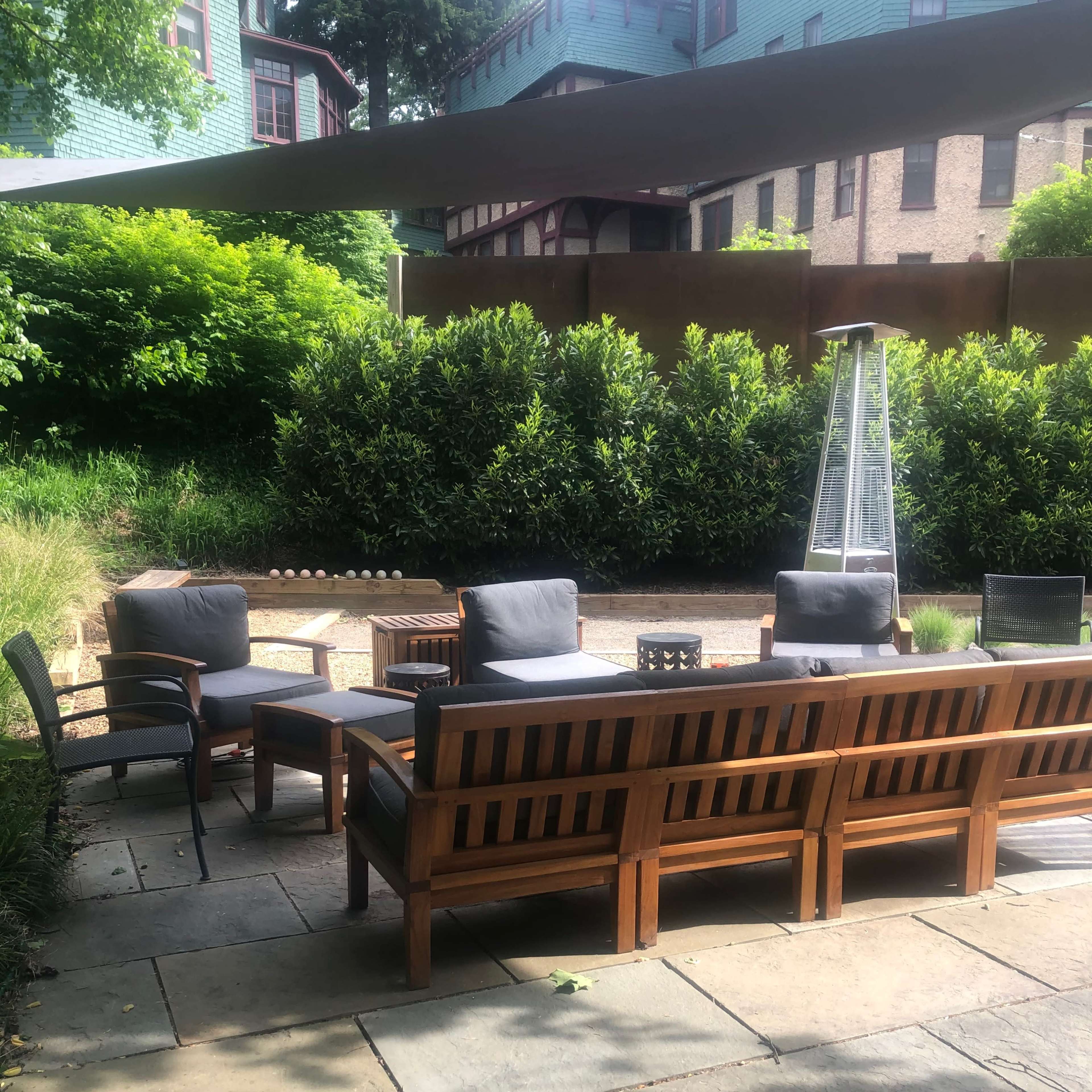 A patio area with wooden furniture arranged around a fire pit, surrounded by greenery and a shaded section.