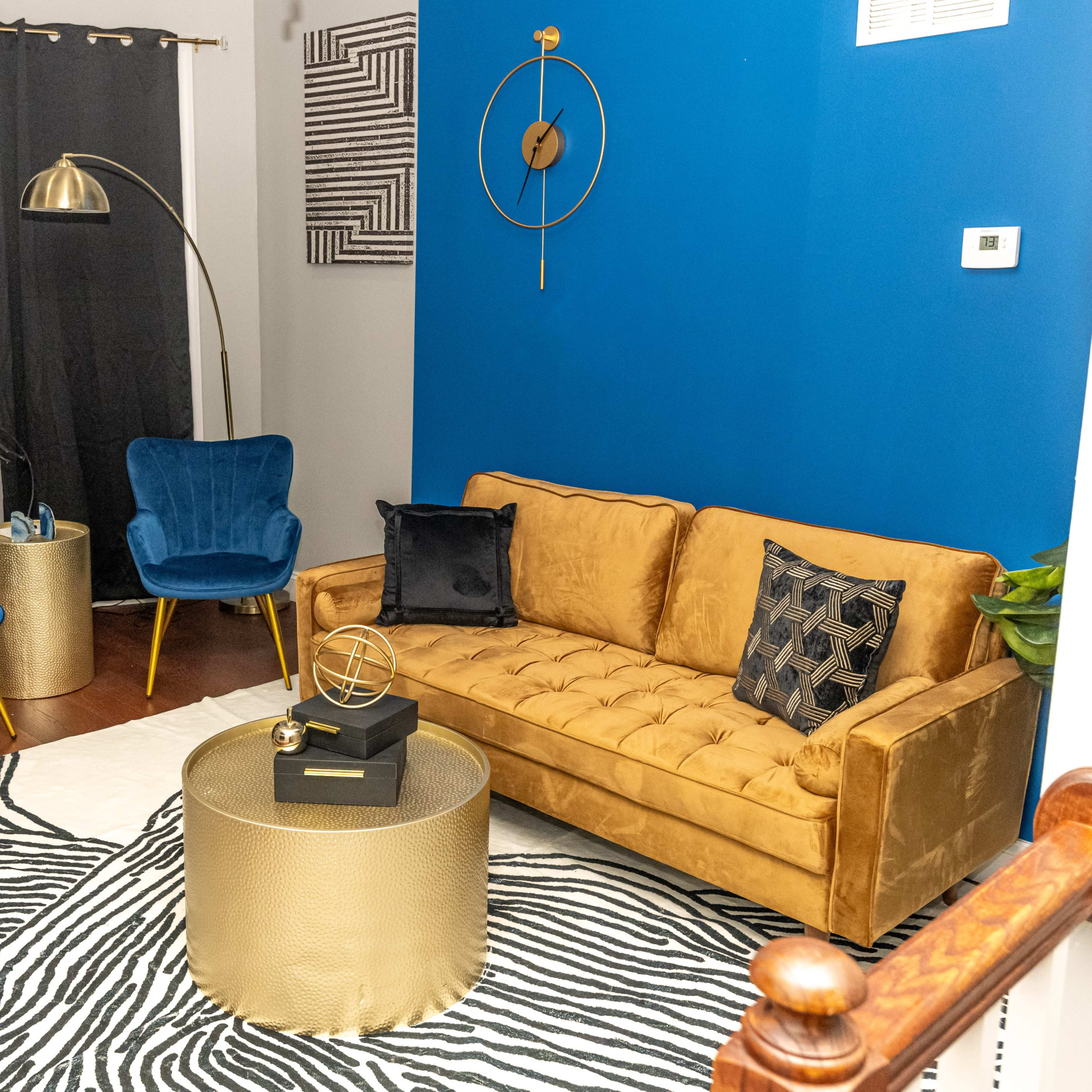 The scene features a cozy living room with a gold tufted sofa, two blue accent chairs, a round gold coffee table, and a zebra-patterned rug against a blue wall.