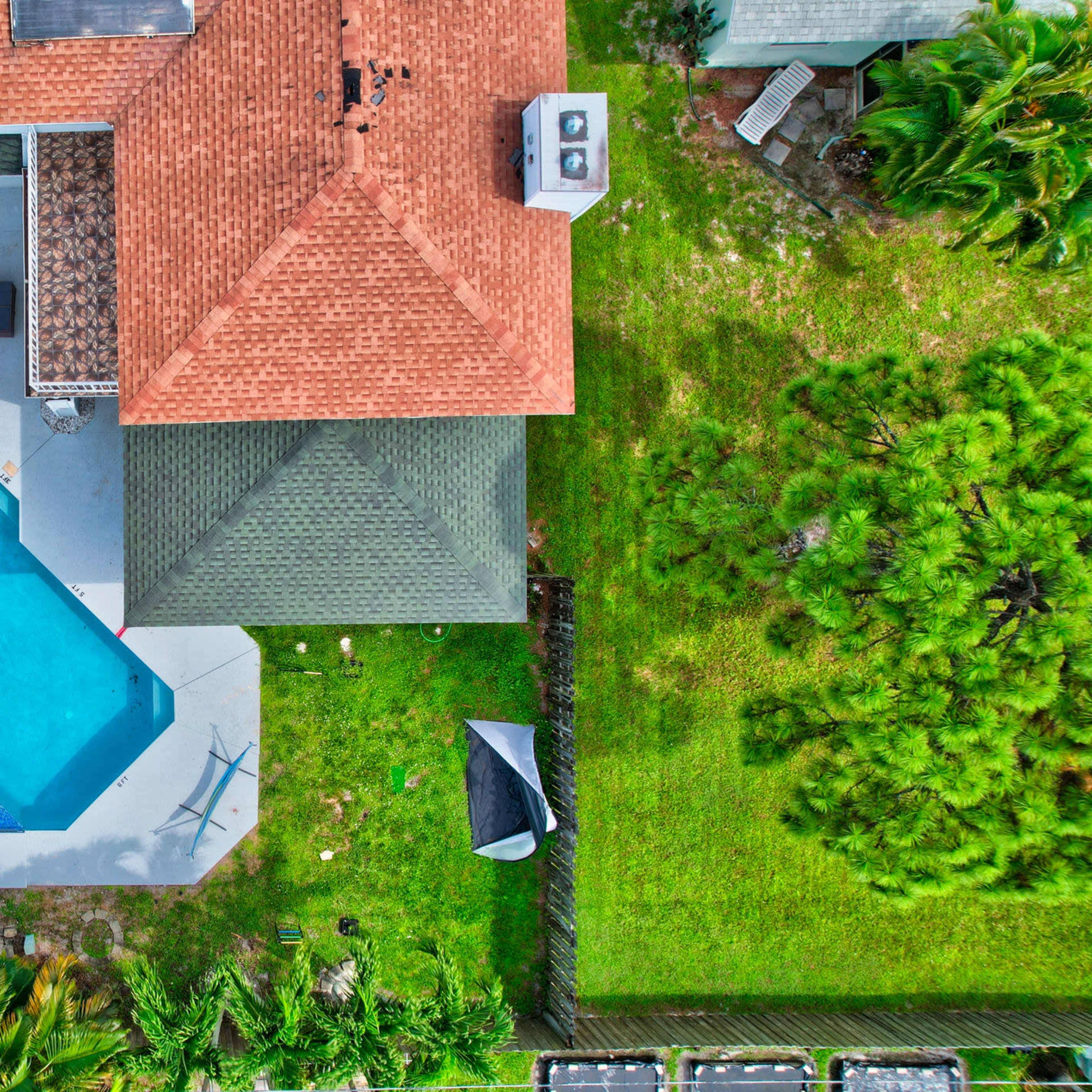 An aerial view shows a backyard with a swimming pool, surrounding patio, and two nearby houses set in green landscaping.