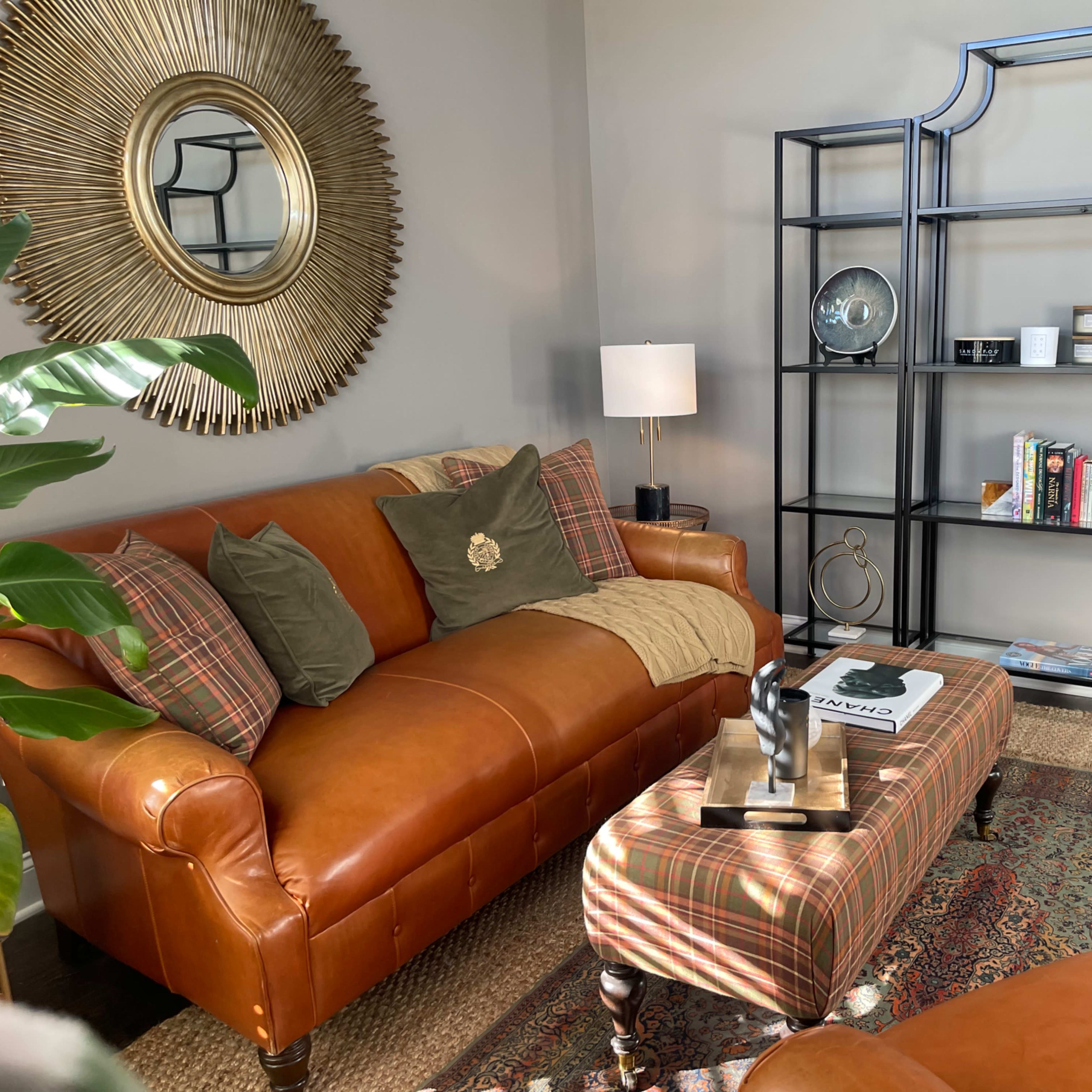 A cozy living room with a brown leather sofa, a patterned ottoman, a bookshelf, and a large circular mirror.