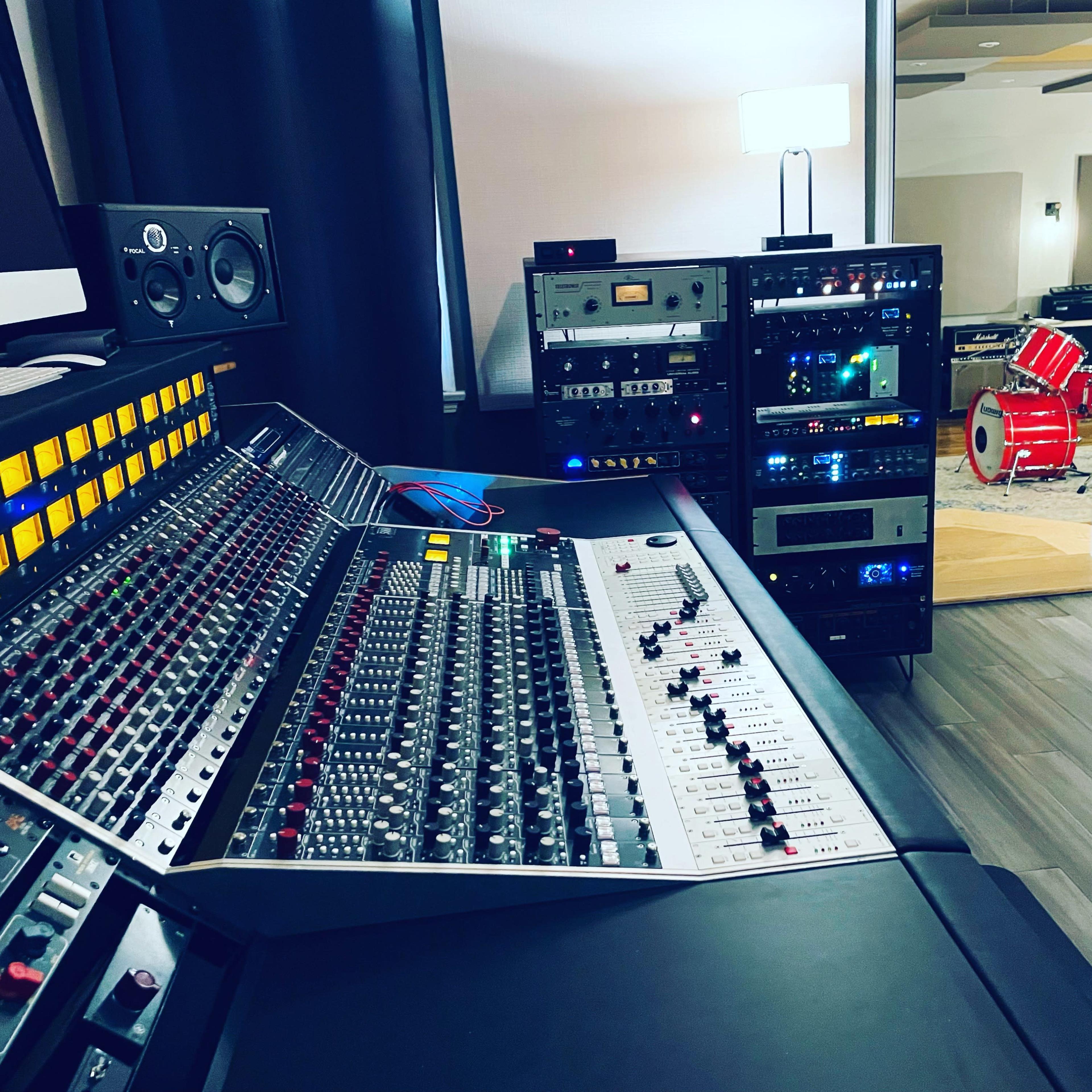 A music studio featuring a large mixing console, various audio equipment racks, and a glimpse of a drum set in an adjacent room.