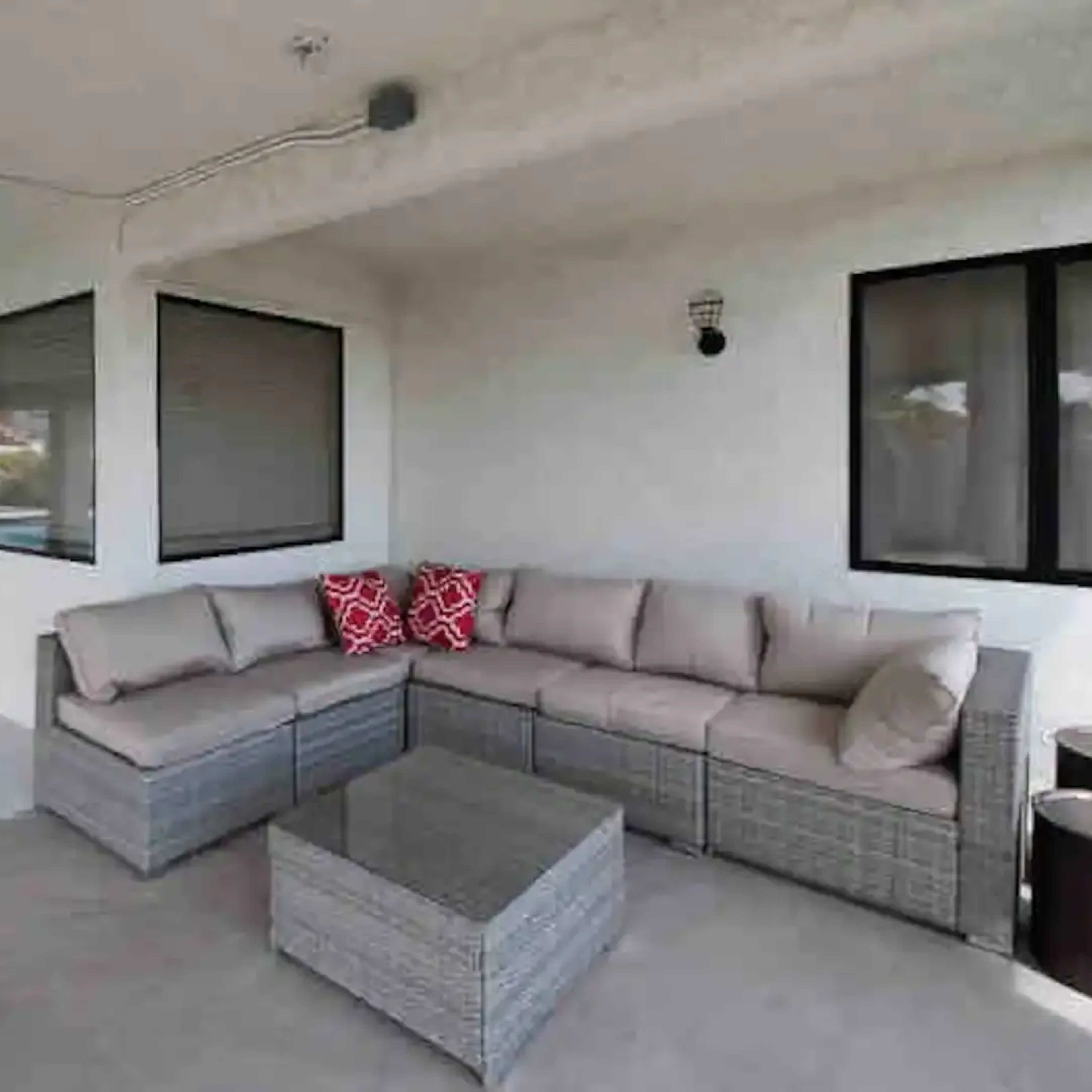 A covered outdoor patio with a sectional sofa and a coffee table, set against white walls and large windows.