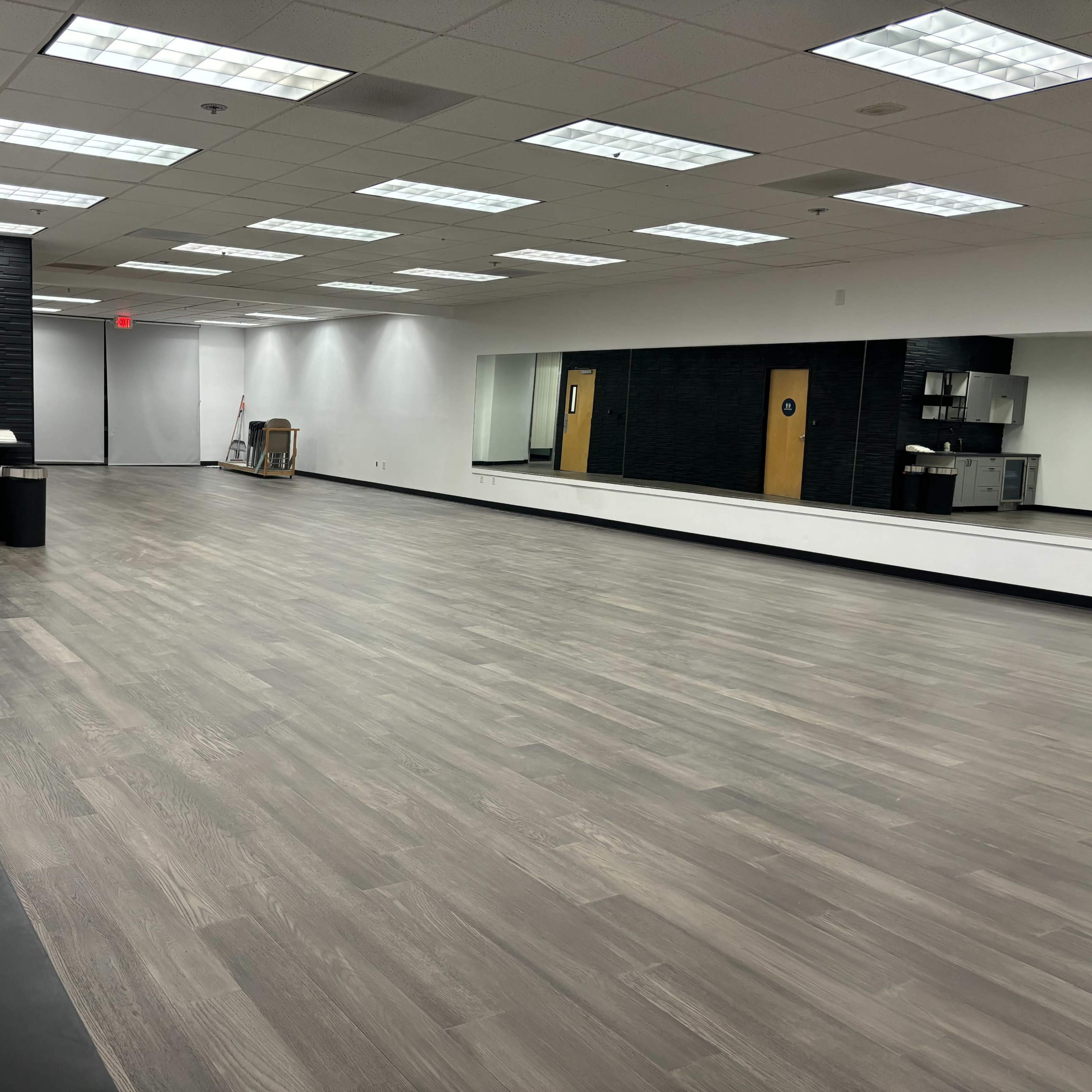 A spacious room with wooden flooring, equipped with a kitchenette on one side and a large mirror along the opposite wall.