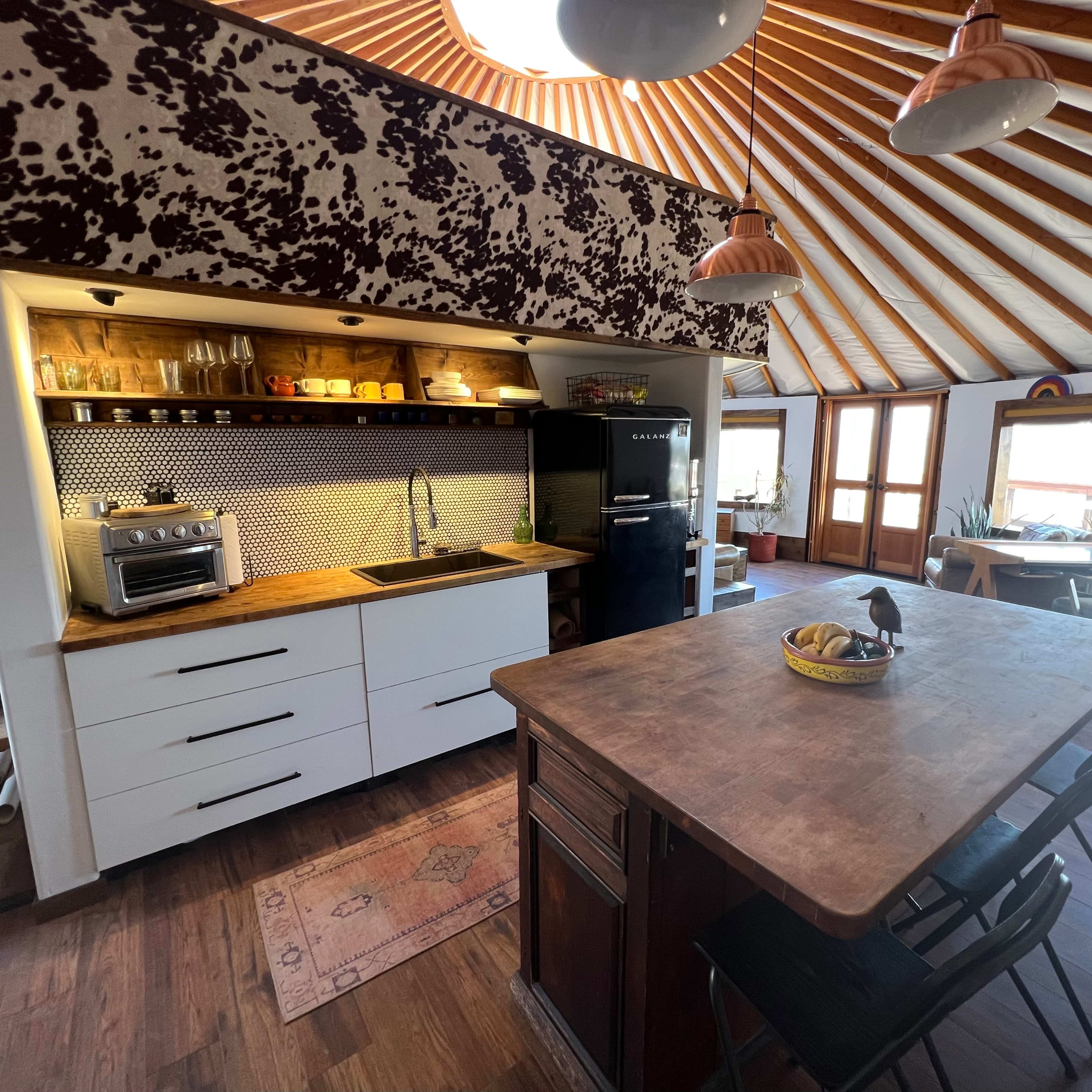 A modern kitchen inside a yurt, featuring a central island with a wooden countertop, a black refrigerator, and large windows allowing natural light.