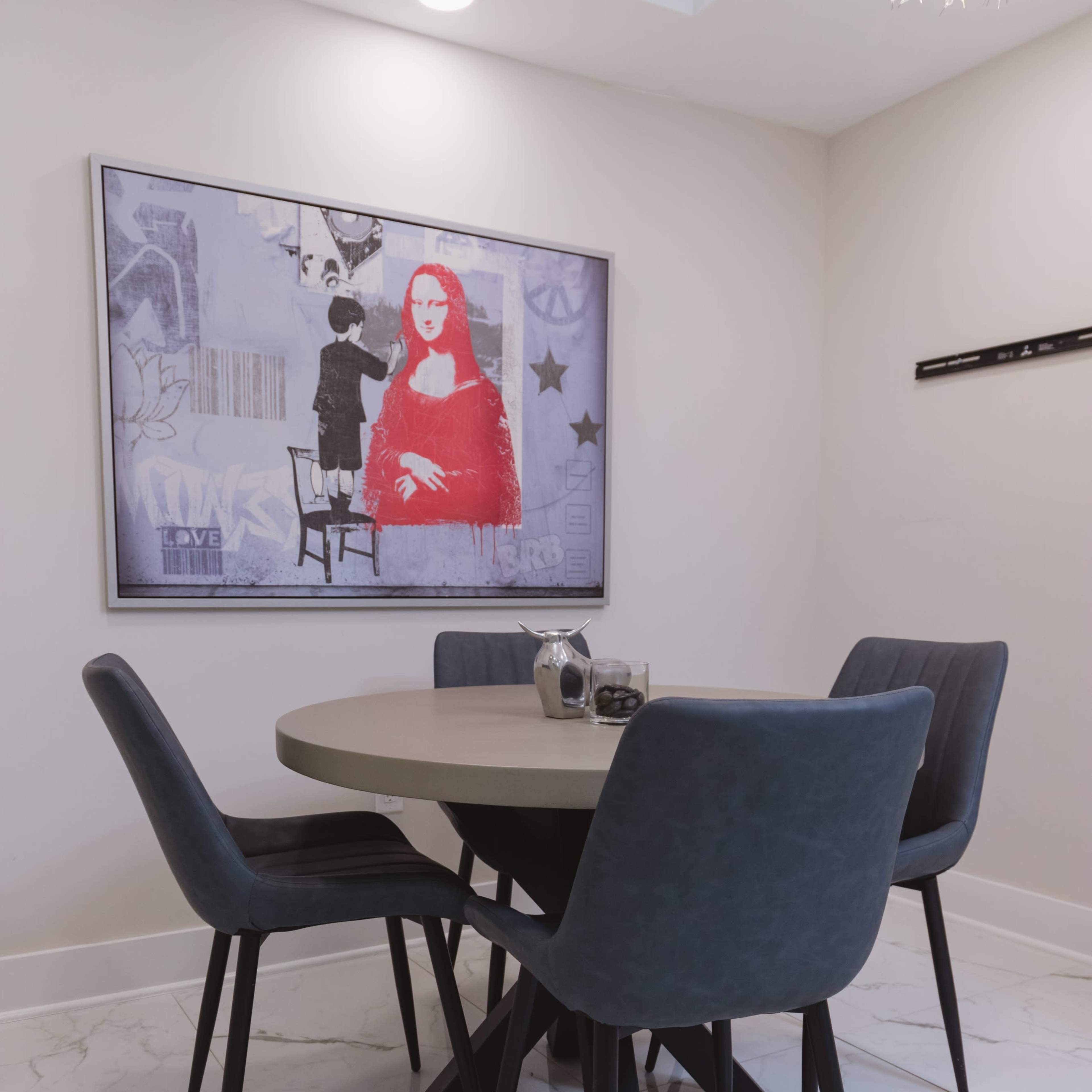 A round dining table surrounded by four upholstered chairs sits in a room featuring a framed artwork on the wall.