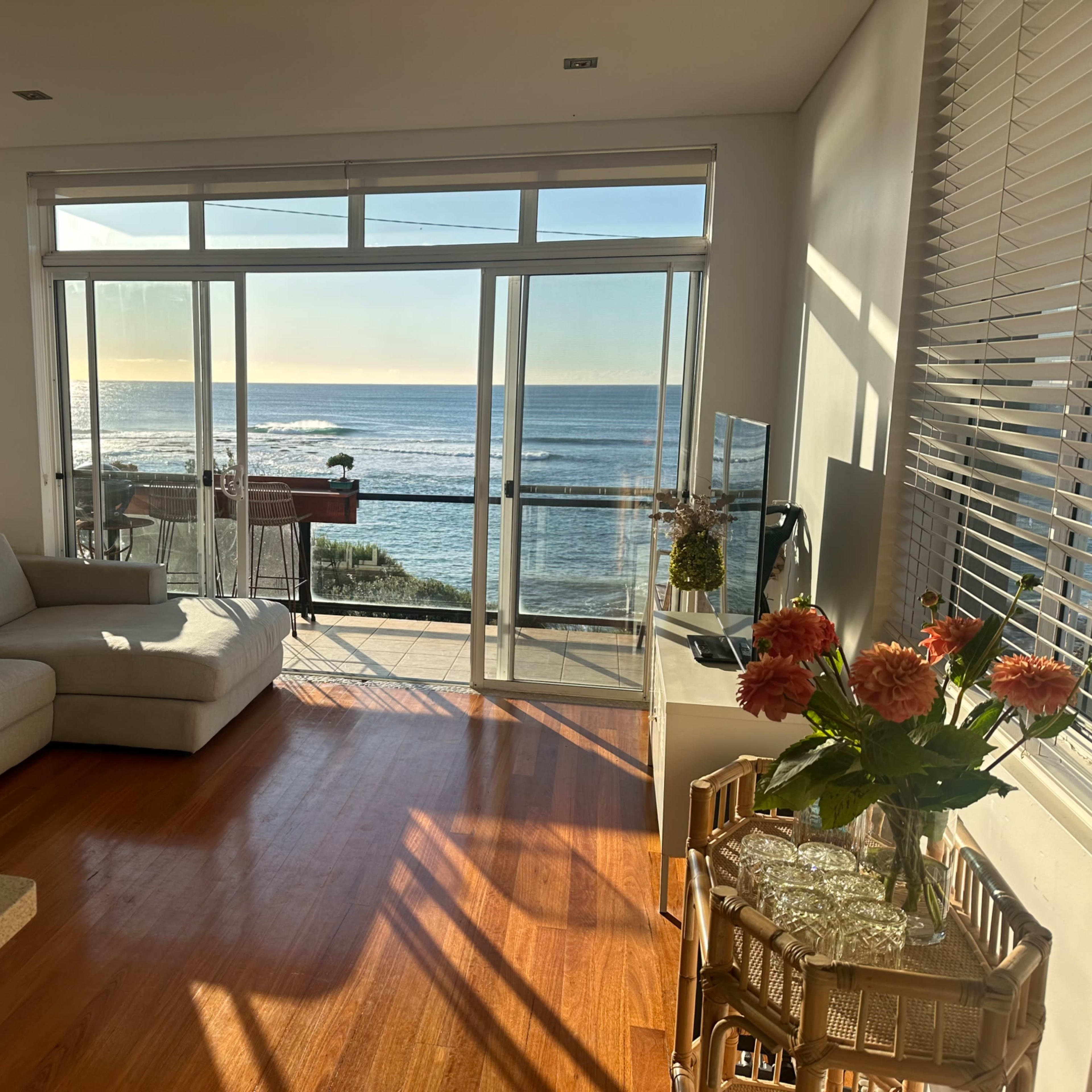 A living room features a gray sofa, a coffee table with flowers, and large windows overlooking the ocean.