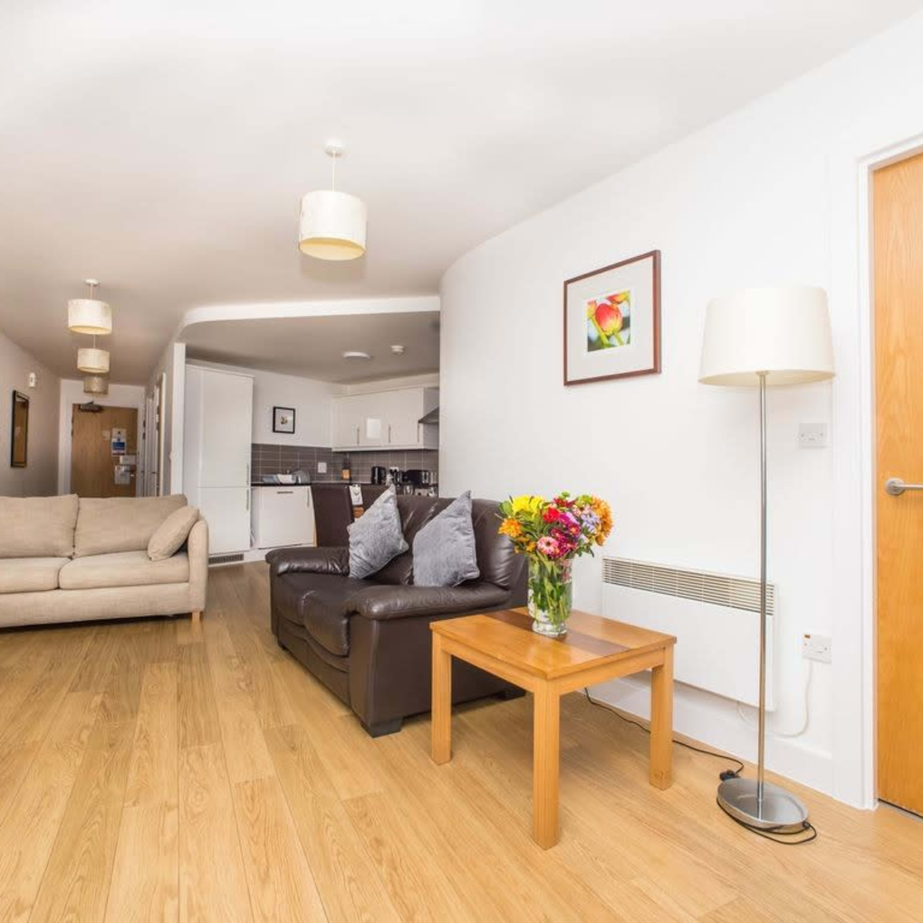 A living room with a brown couch, a wooden coffee table with a vase of flowers, and a light-colored wall leading to a kitchen area in the background.