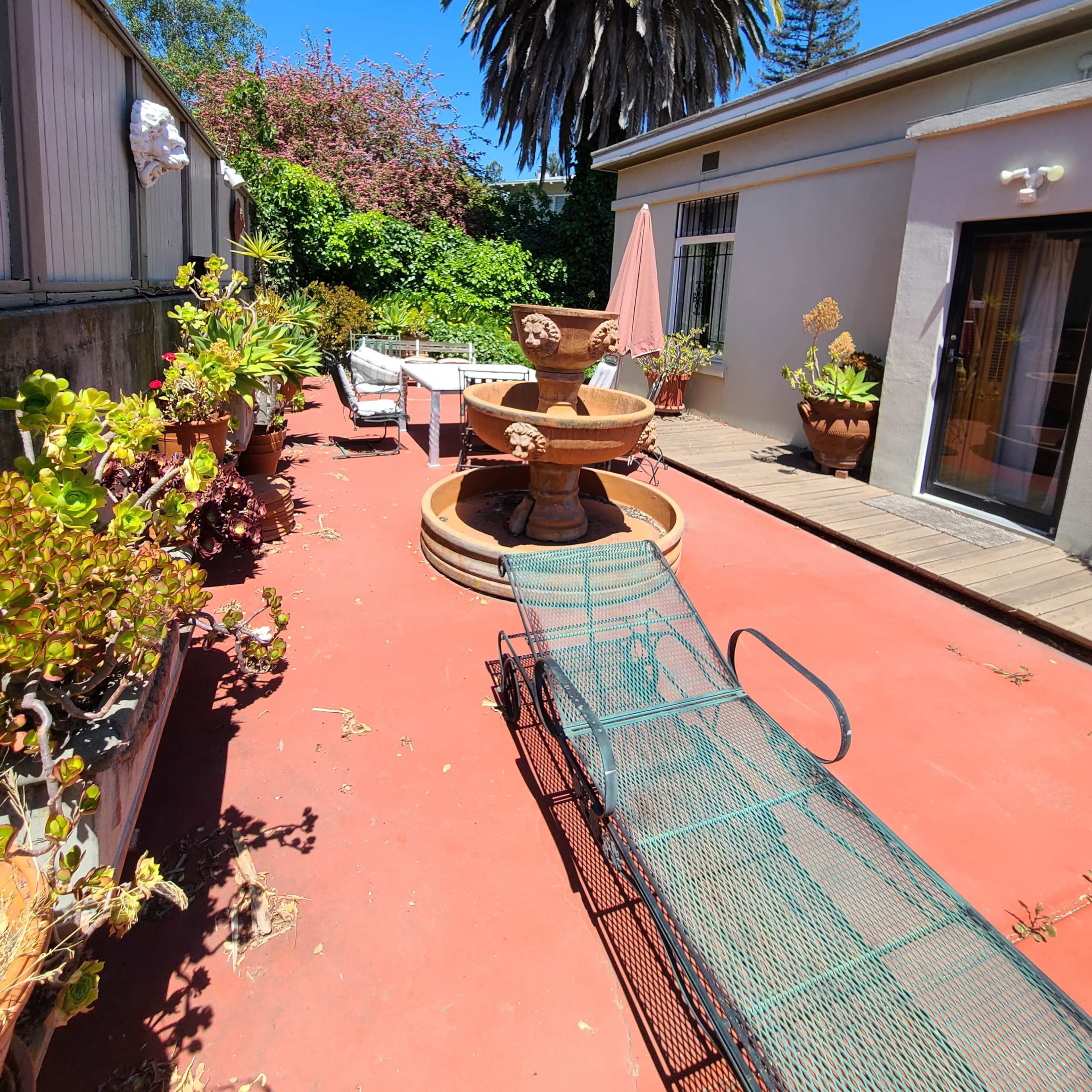 A patio area featuring a circular fountain surrounded by potted plants and a lounge chair on a red surface, with a tall palm tree in the background.