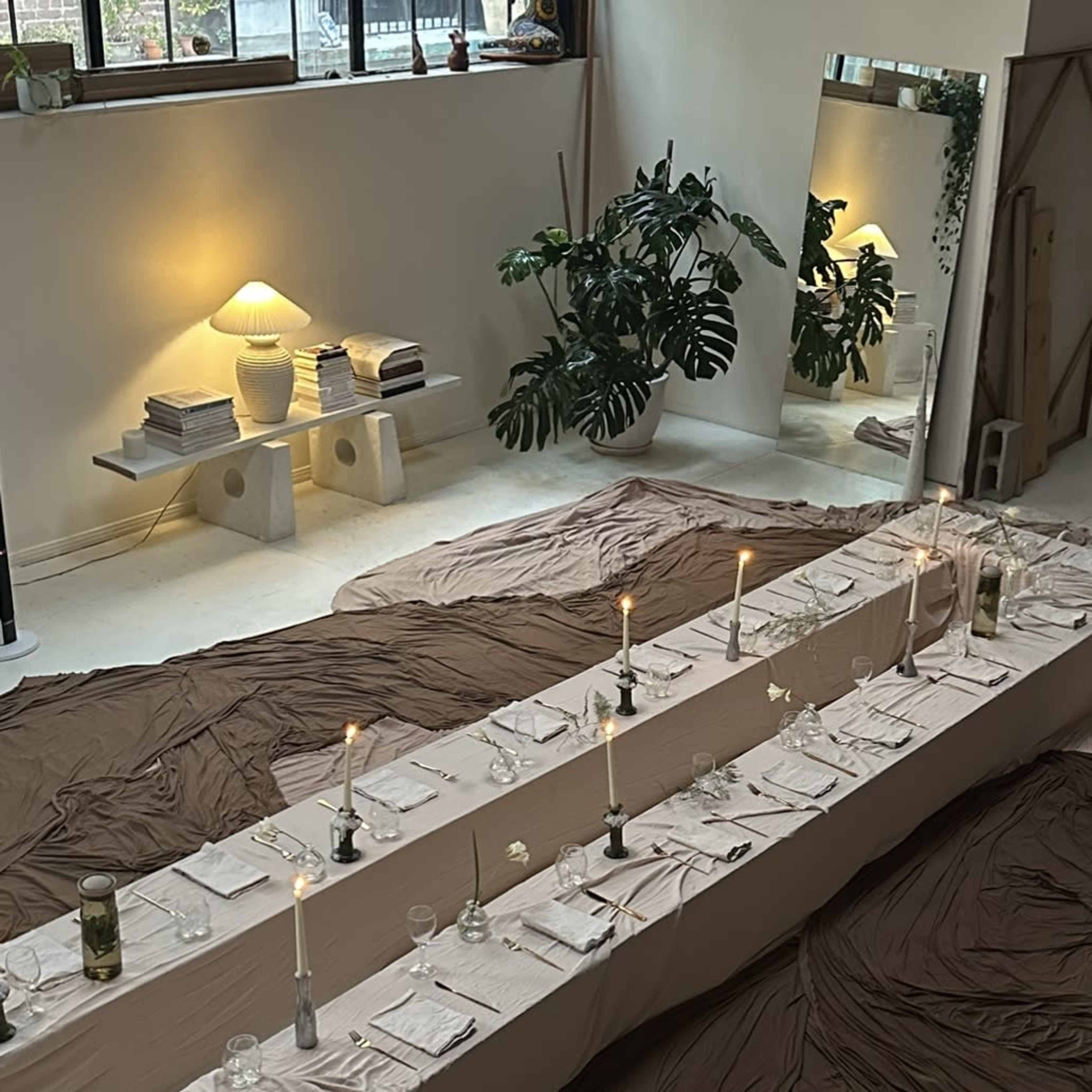 A long dining table is set with plates and candles, surrounded by draped fabric and plants in a bright room.