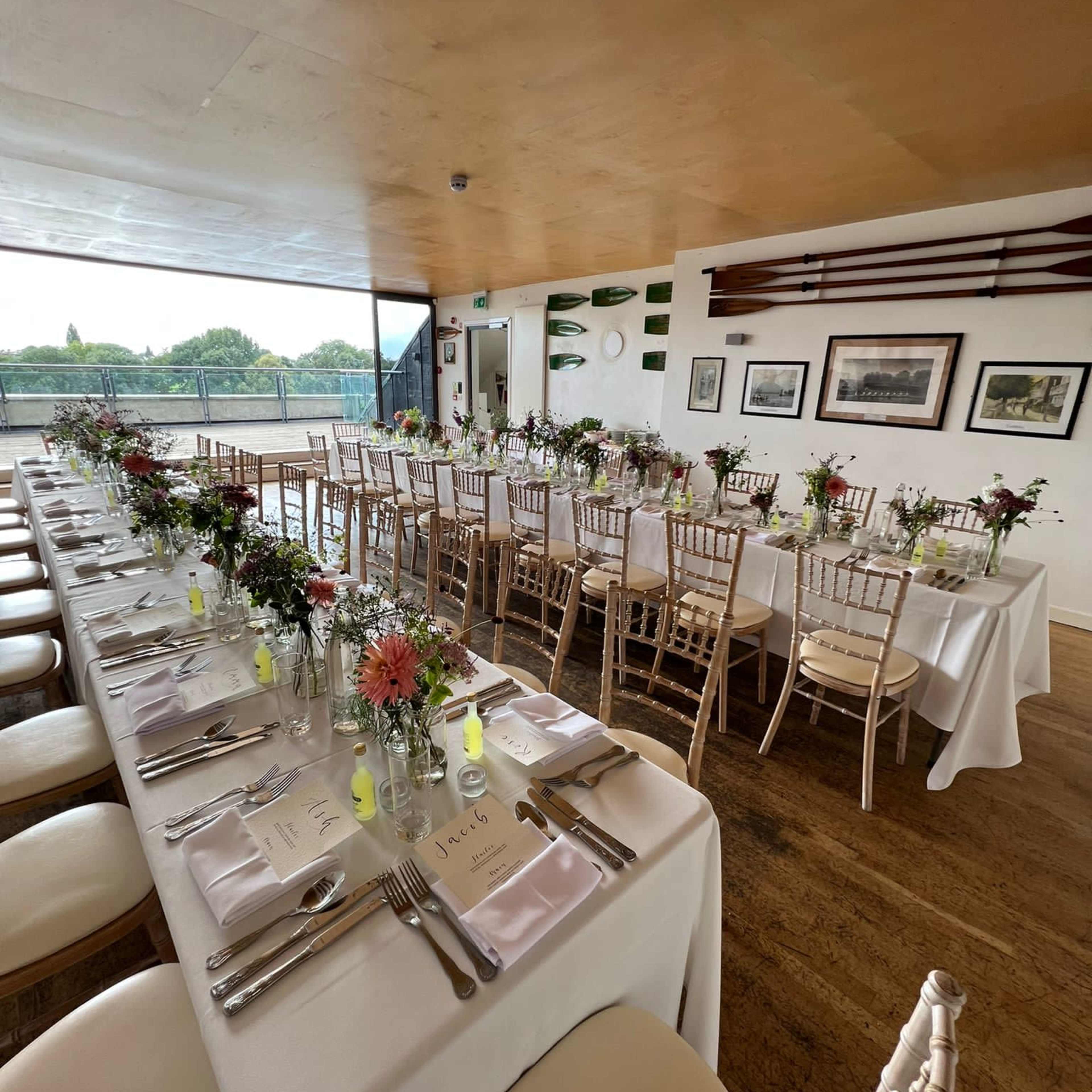 A long dining table is set with white tablecloths and floral centerpieces in a modern room overlooking a green landscape.