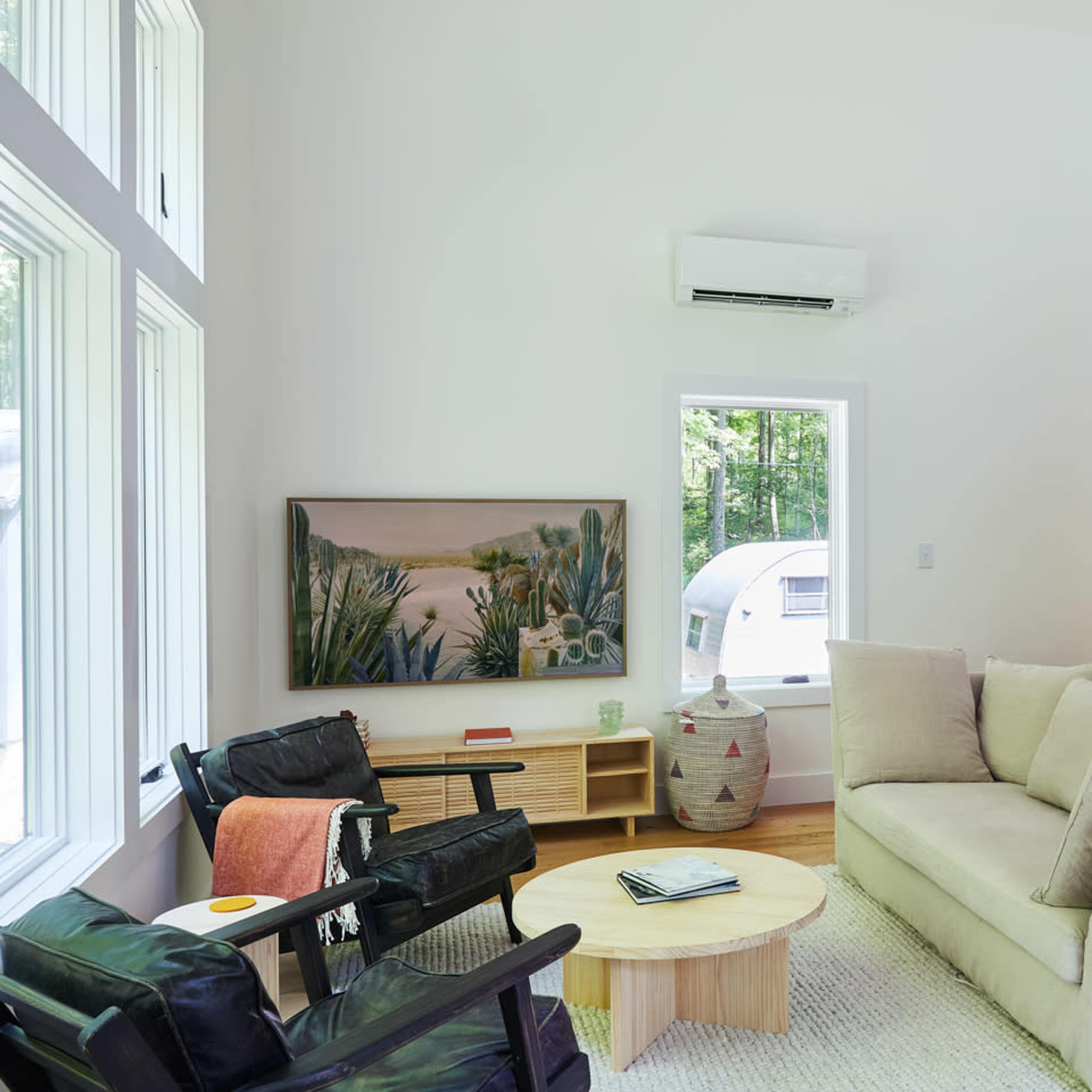 A modern living room with a beige sofa, black armchairs, a wooden coffee table, and a staircase with metal railings, all placed next to large windows allowing natural light.