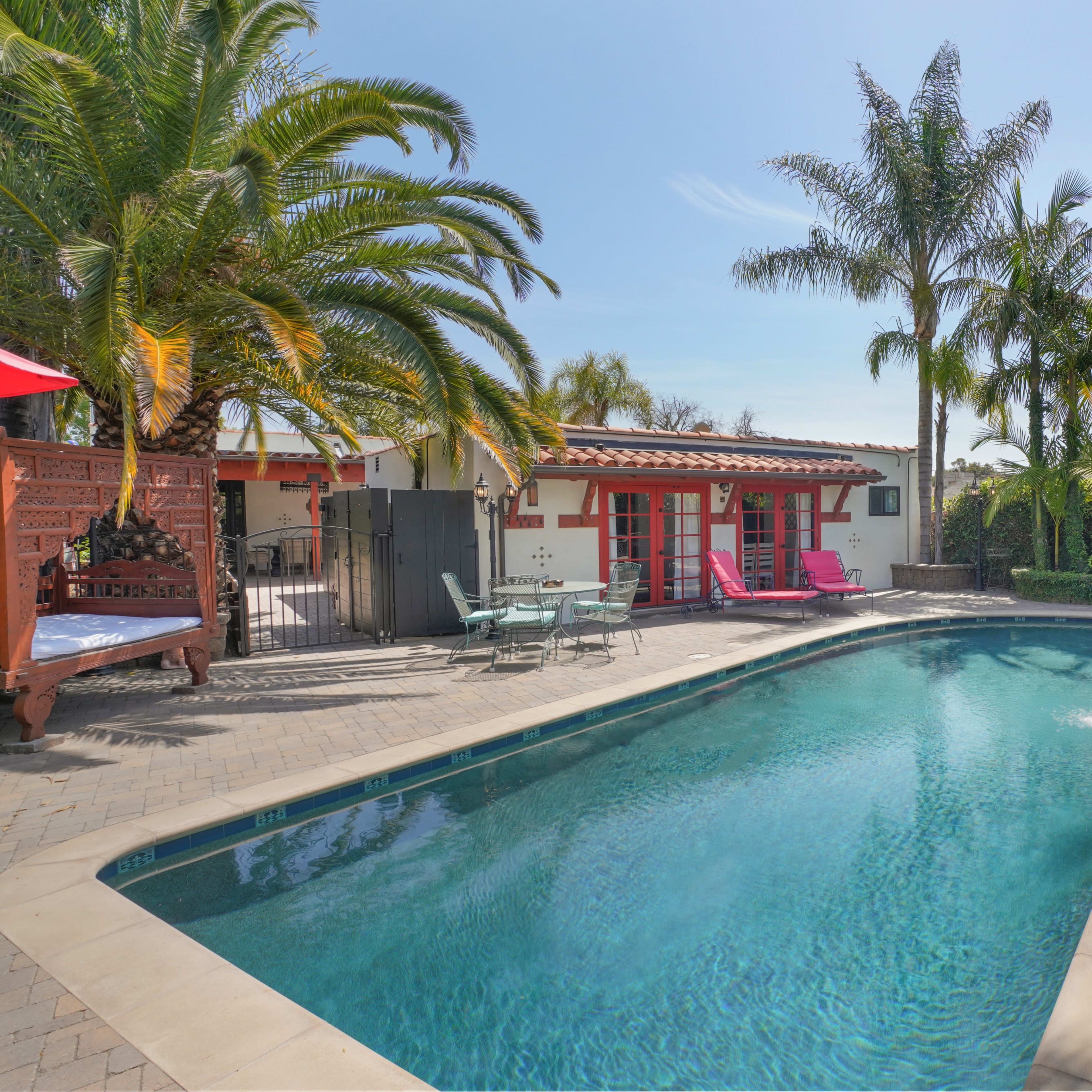 A swimming pool surrounded by palm trees and outdoor seating in a sunlit backyard.