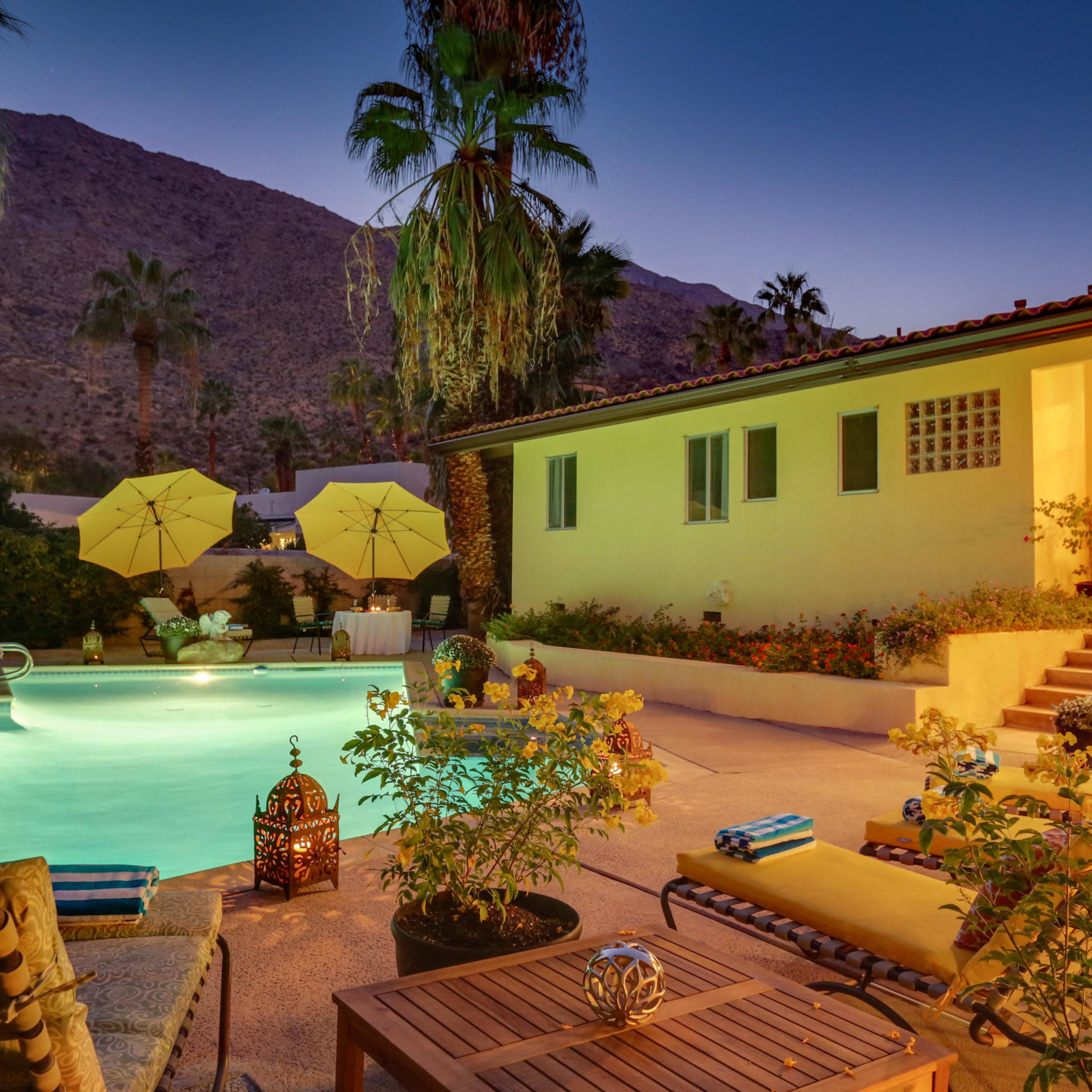 A well-lit backyard features a swimming pool surrounded by lounge chairs, potted plants, and lanterns, with a house and mountains in the background at dusk.