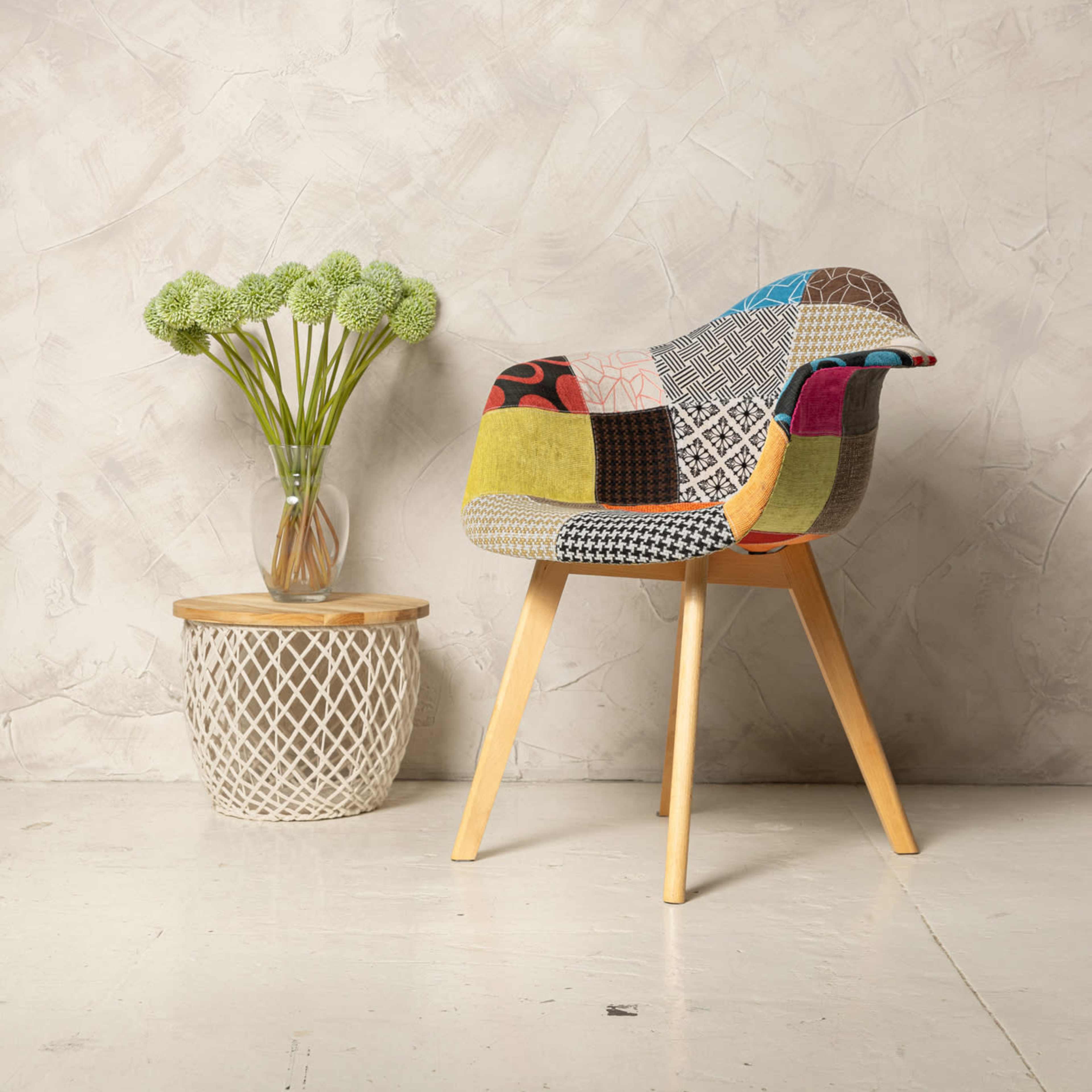 A colorful patchwork chair sits beside a braided basket table holding a vase of green flowers against a textured grey wall.