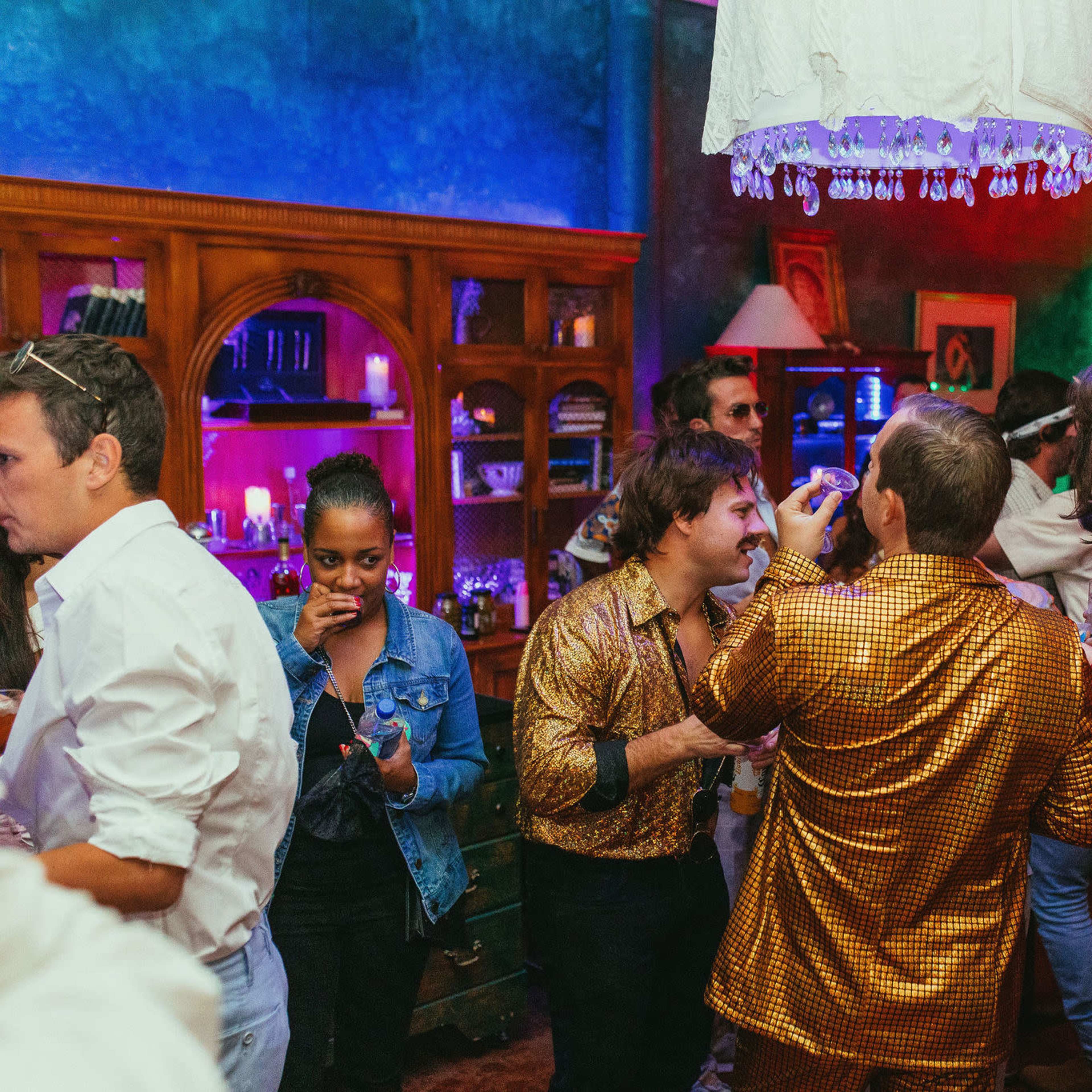 A lively party scene features guests in colorful outfits mingling and enjoying drinks in a vibrant setting with eclectic decor.