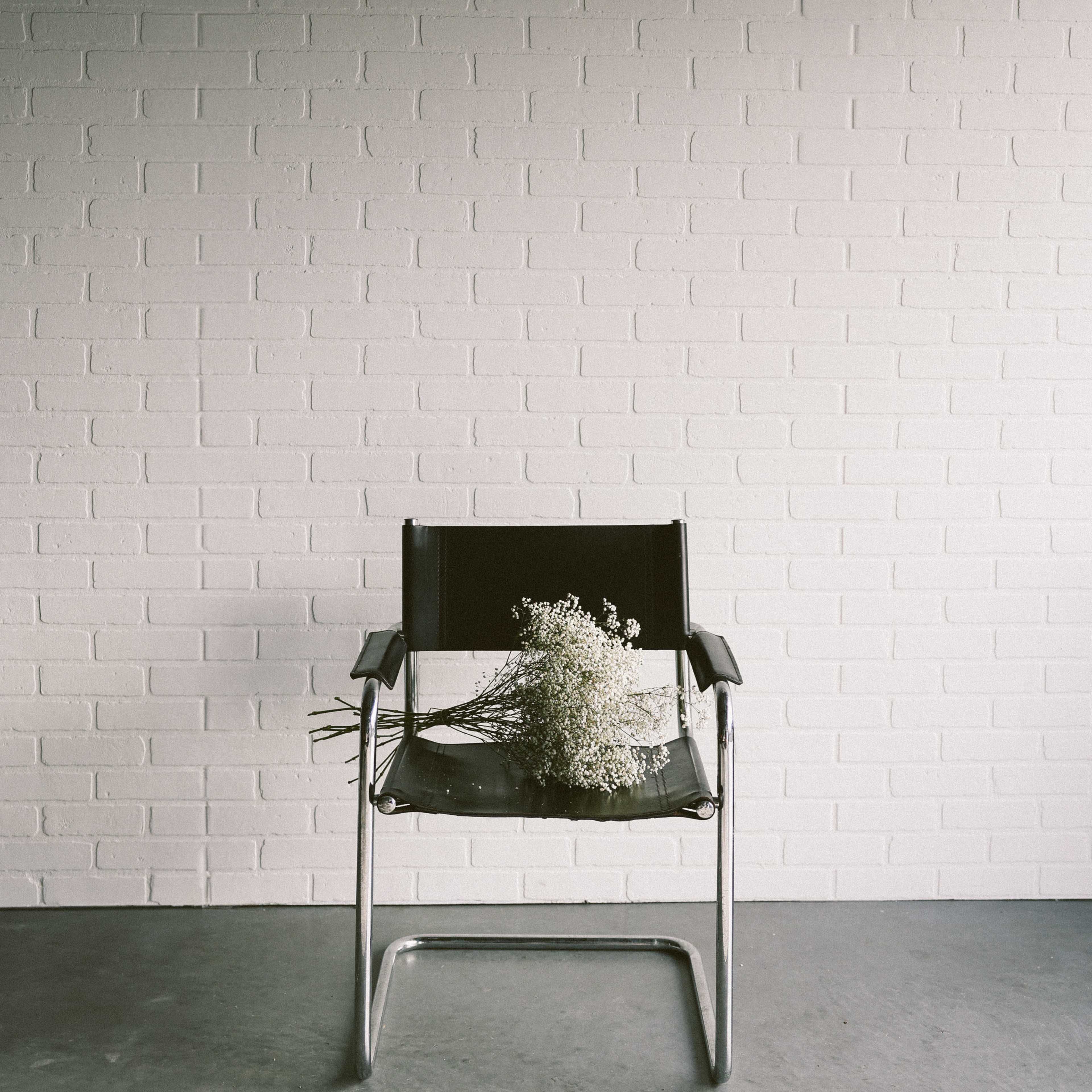 A black chair with metal legs sits against a white brick wall, adorned with a bouquet of dried flowers.