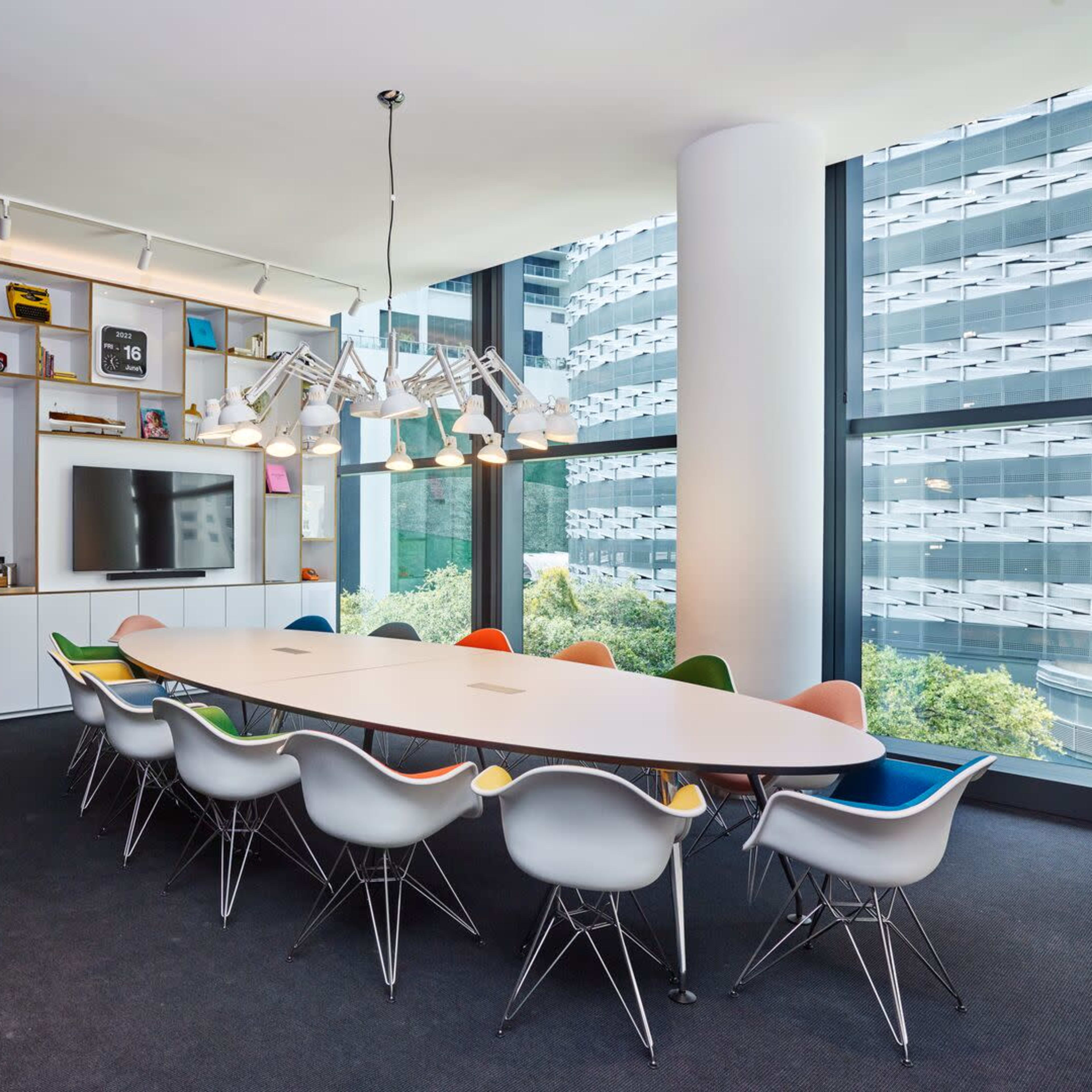 A spacious conference room features a long oval table surrounded by colorful chairs, with large windows offering a view of the outside.