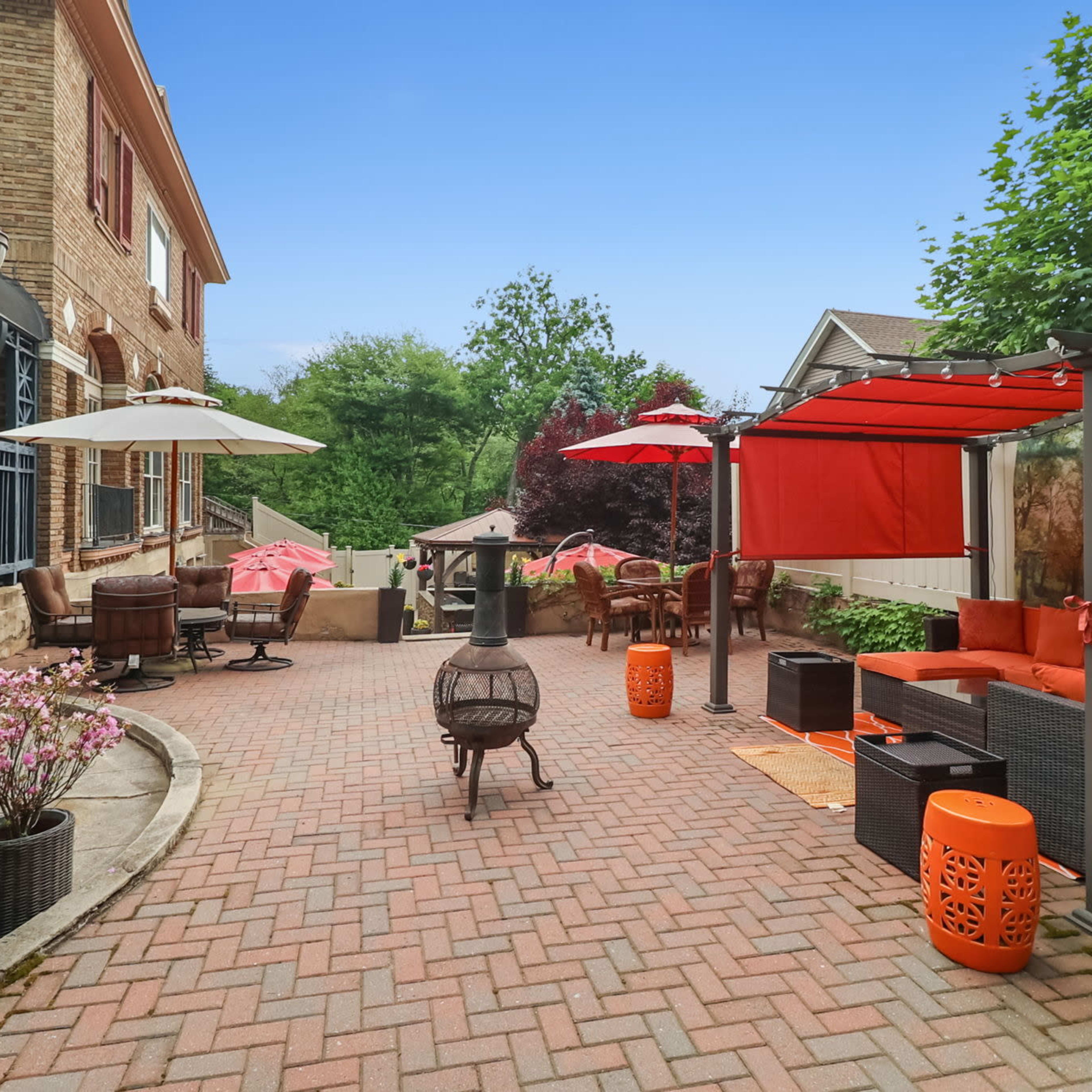 A brick patio features various seating areas with umbrellas and a fire pit, surrounded by greenery and decorative plants.