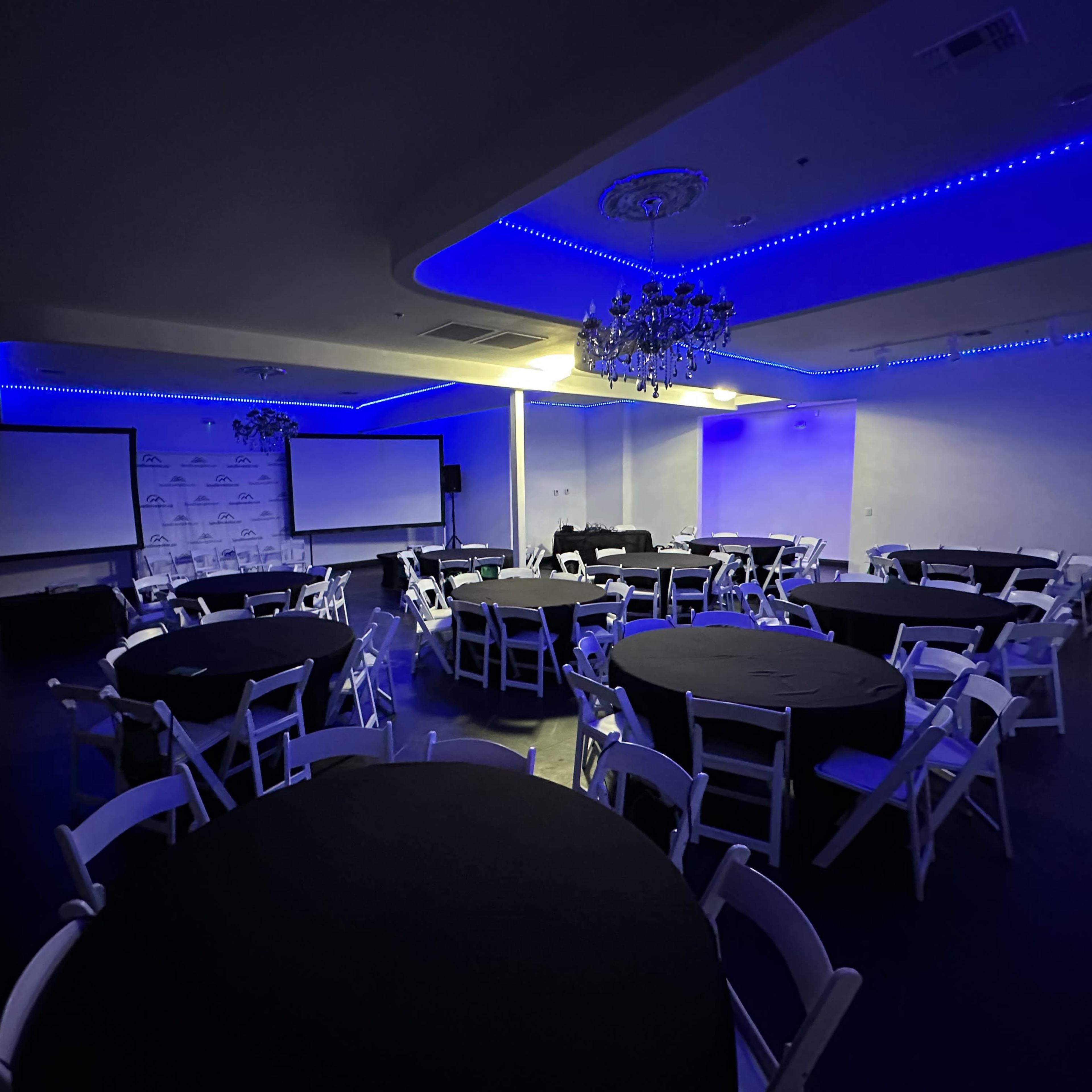 A well-lit event space features round tables with black covers and white chairs arranged around the room, accented by blue LED lighting and two projection screens.