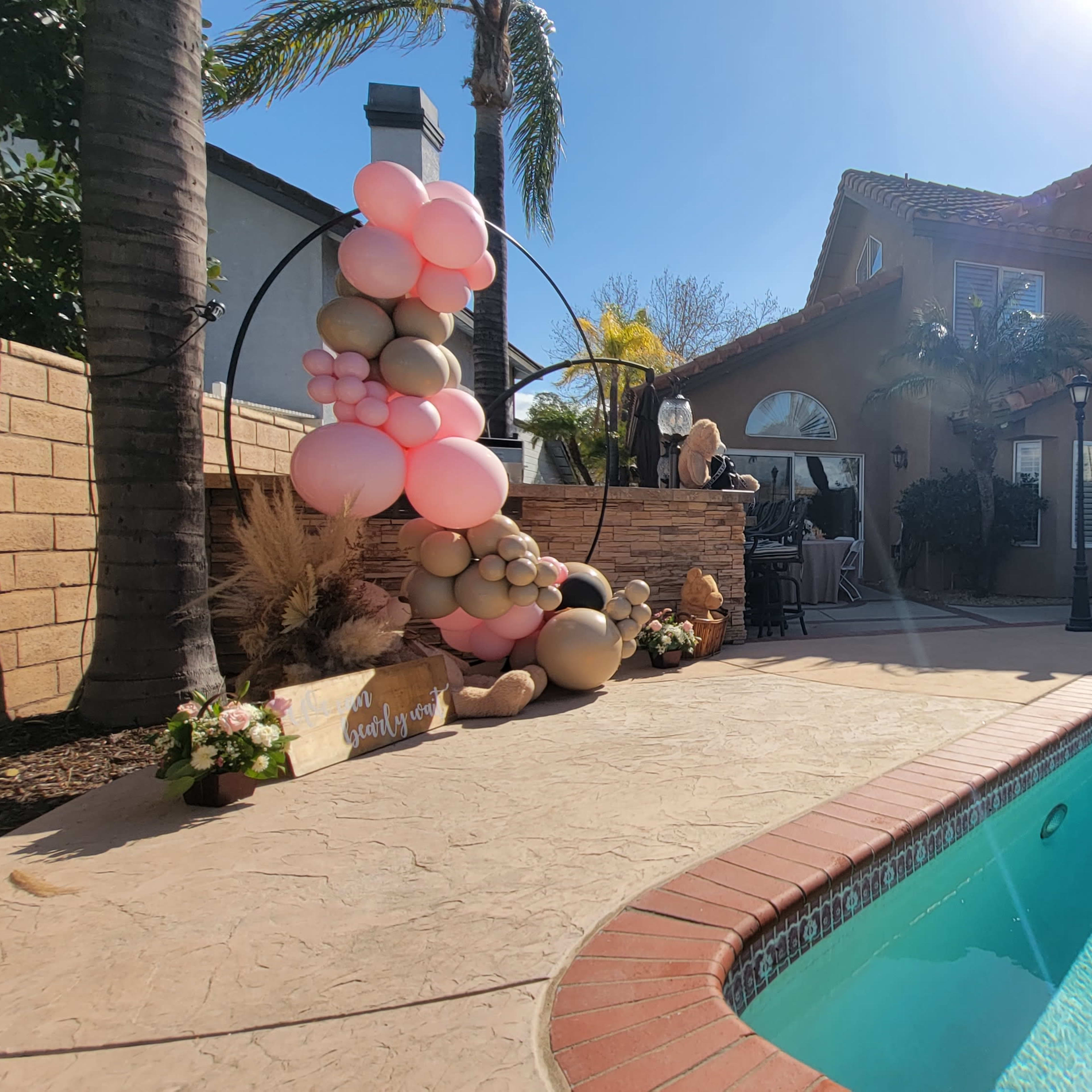A poolside scene features a decorated area with a balloon arch, a patio with seating, and a house in the background under clear blue skies.