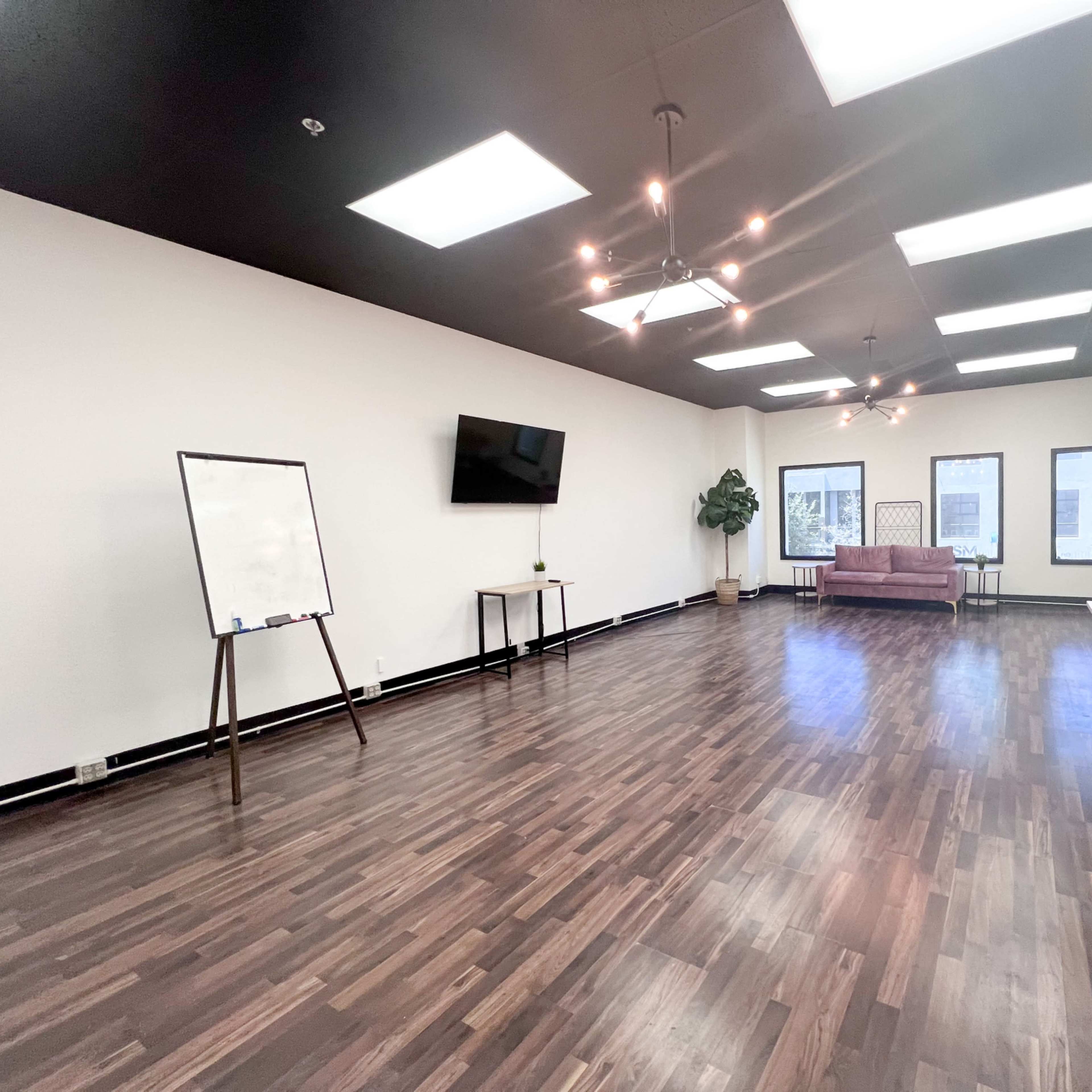 A spacious interior room features a large wooden floor, a television on the wall, a whiteboard stand, a couch, and several windows.