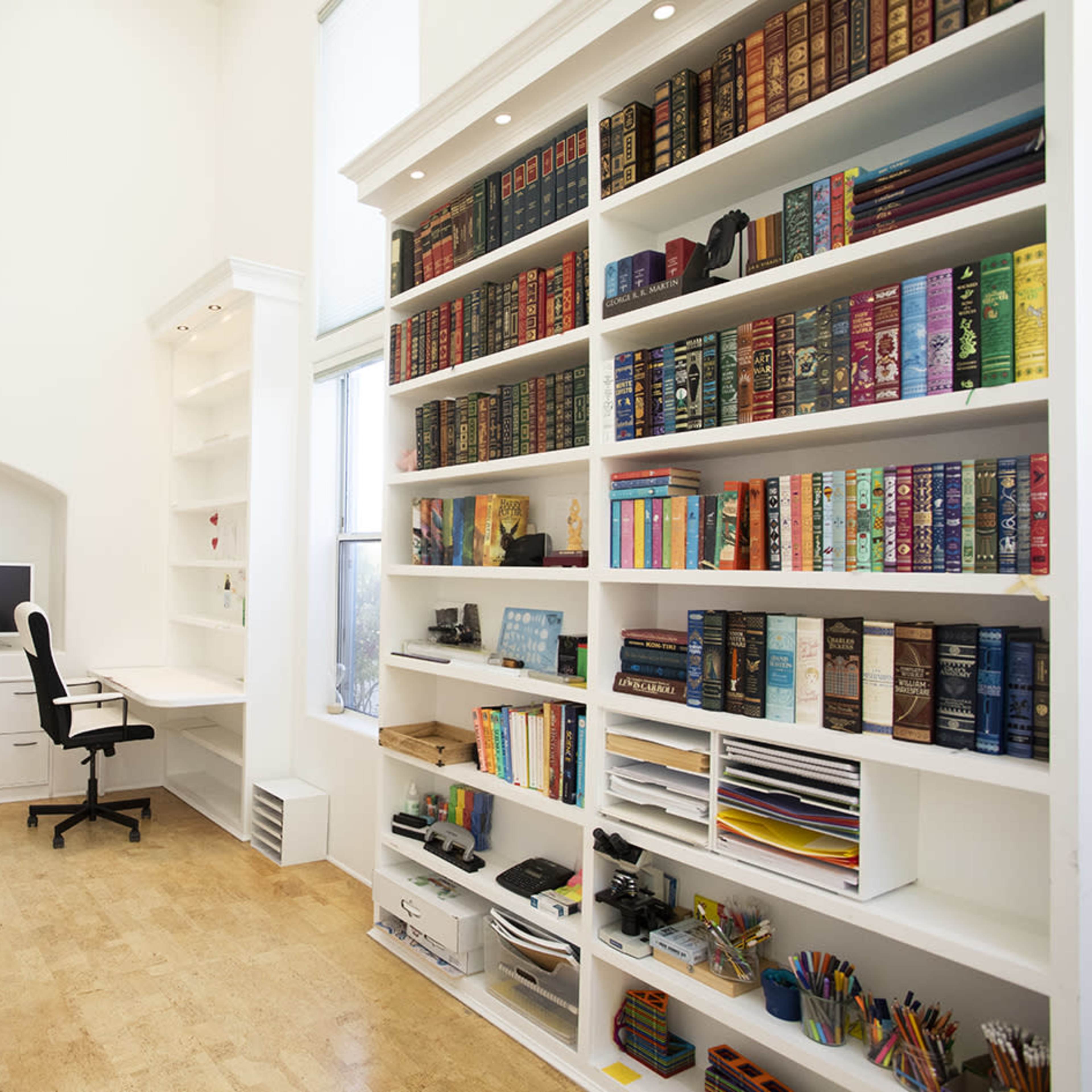 A well-organized home office features white shelves filled with books and a workspace with a desk, chair, and computer.
