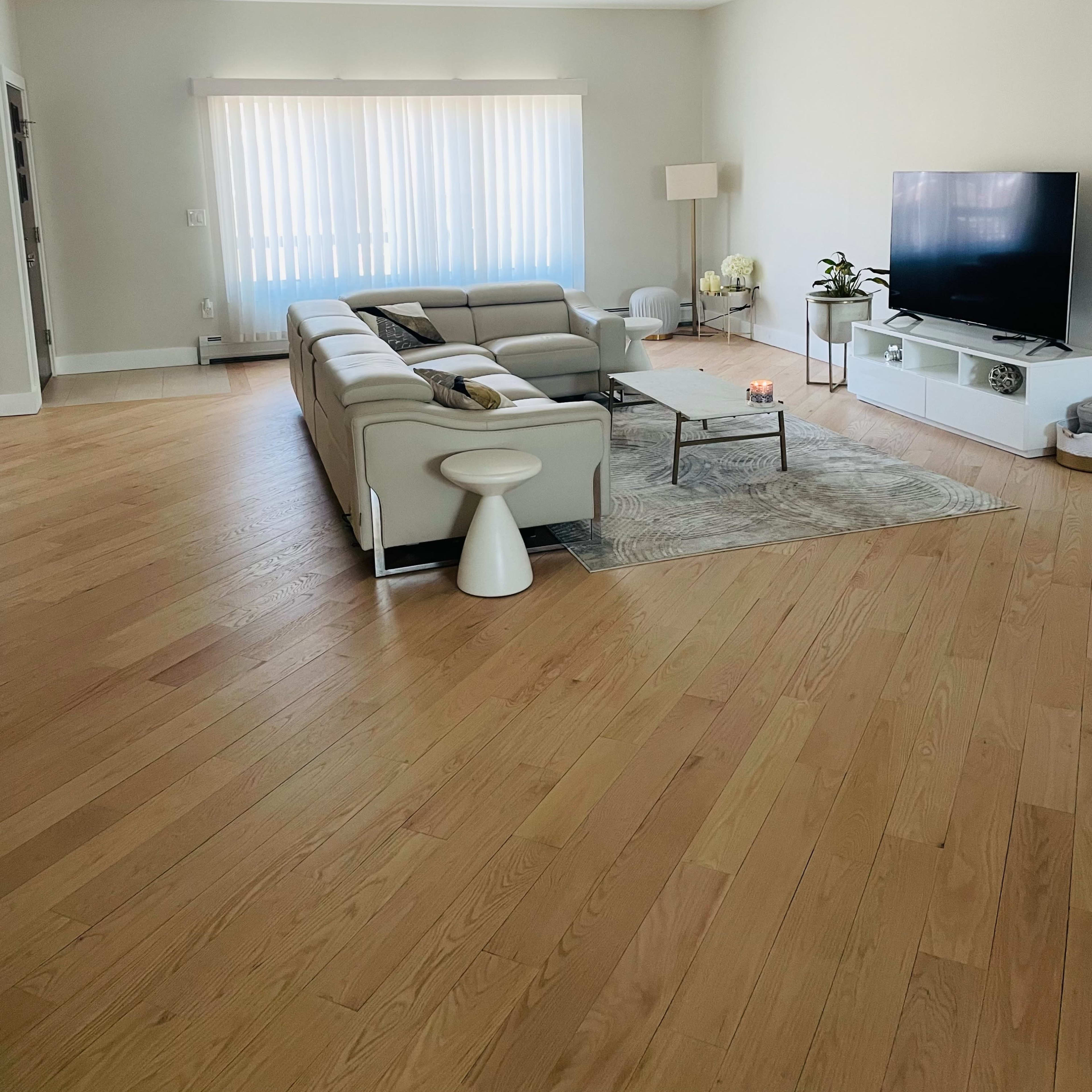 A modern living room features a light-colored sectional sofa, a coffee table, a floor lamp, and a television against a wall with large windows partially covered by sheer blinds.