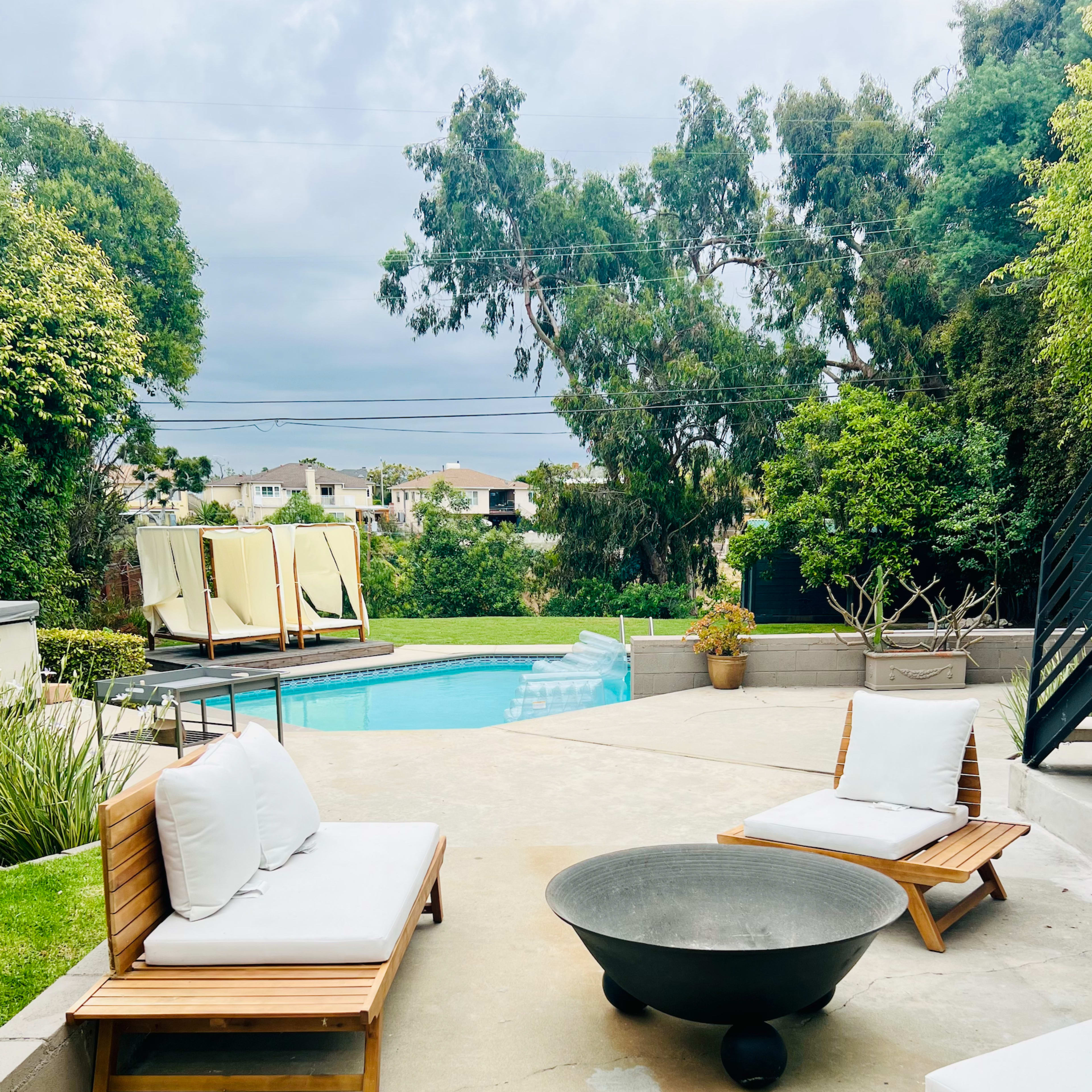 The scene features a sunny backyard with a swimming pool, two lounge chairs, a fire pit, and lush greenery in the background.