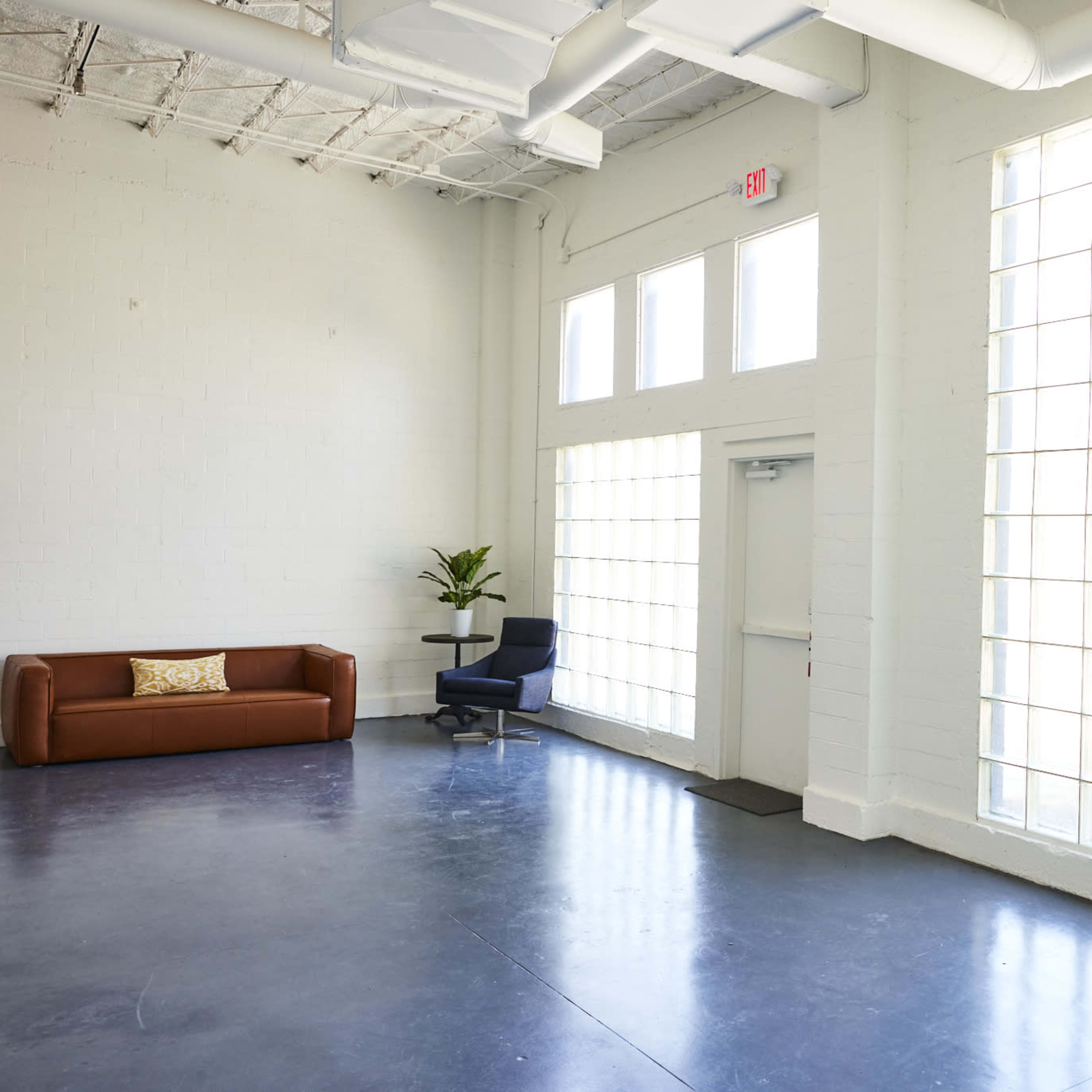 A spacious, minimally furnished room featuring a brown sofa, a blue chair, and large windows allowing ample natural light.