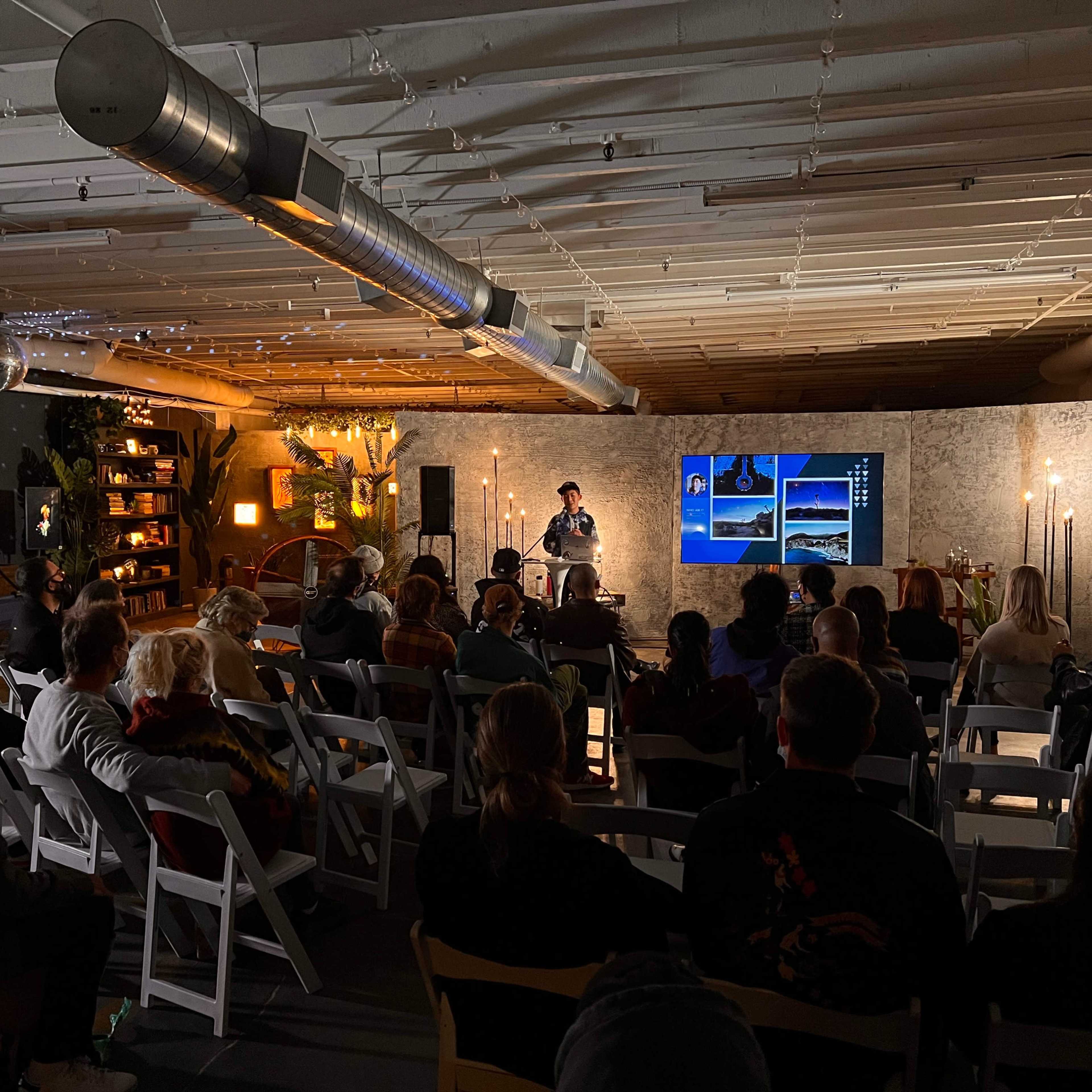 A speaker presents to an audience seated in a well-lit room adorned with plants and modern decor.