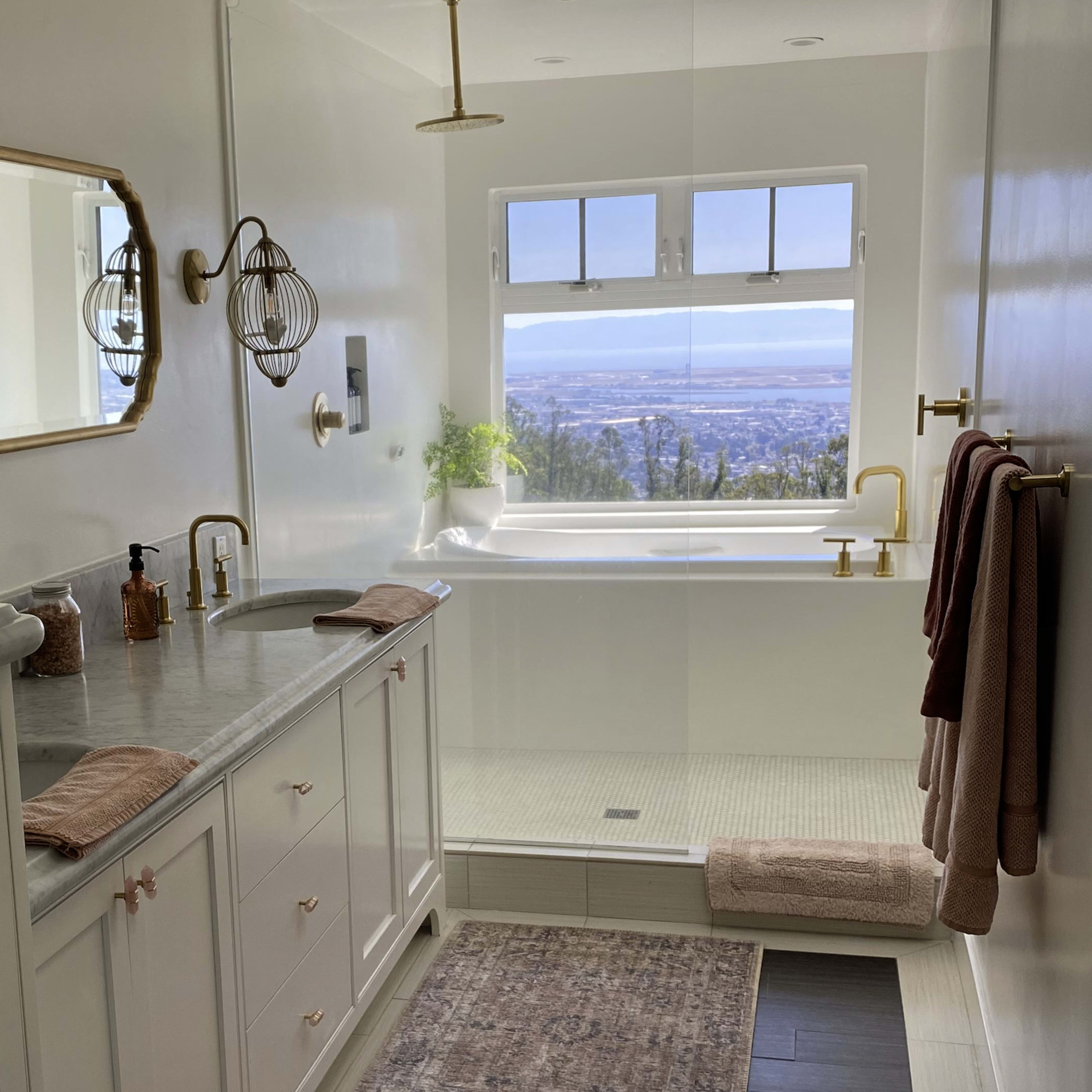 A modern bathroom features a double vanity with a marble countertop, a large window overlooking a view, a freestanding bathtub, and beige towels hung nearby.