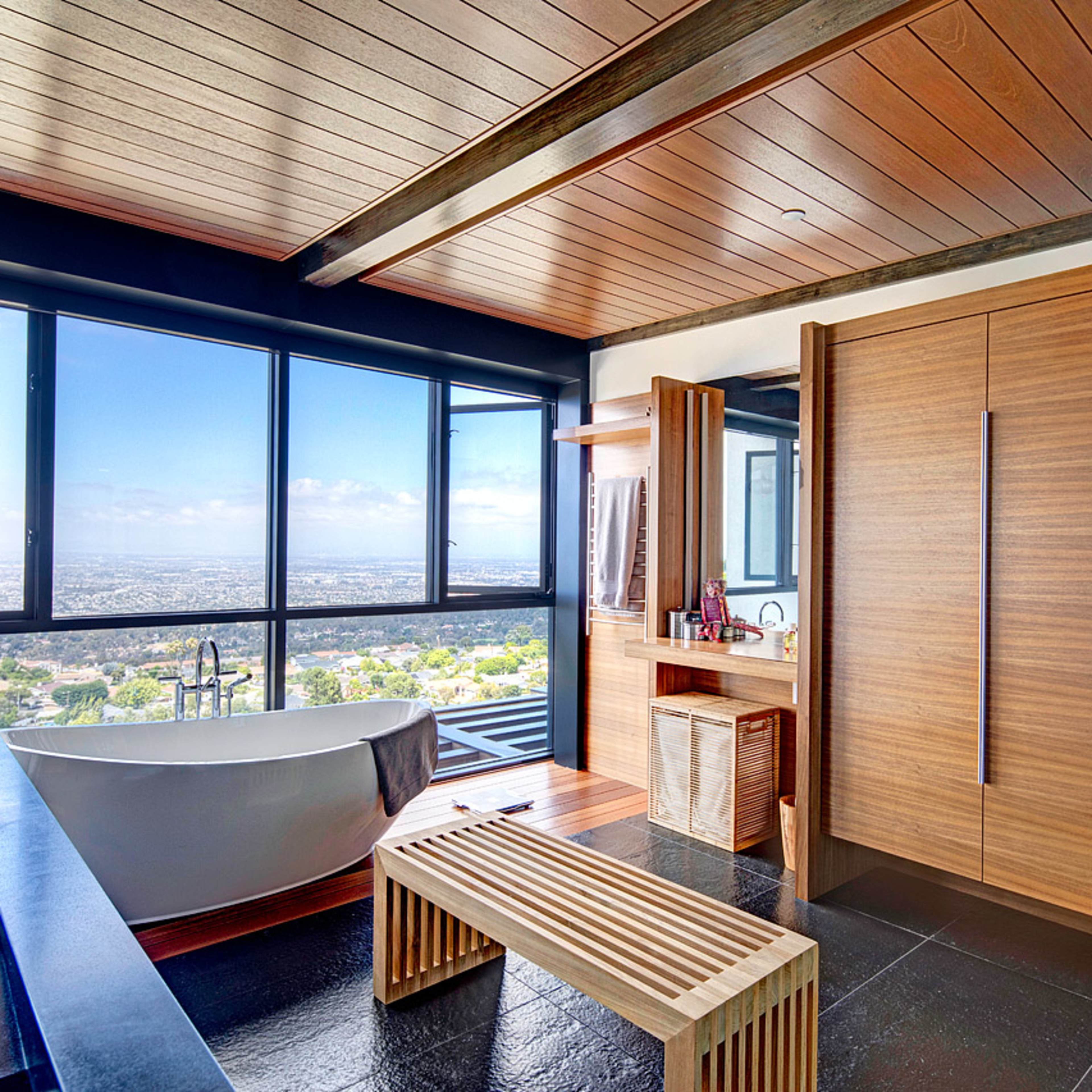 A modern bathroom features a large soaking tub, wooden cabinetry, and expansive windows offering a view of the landscape outside.