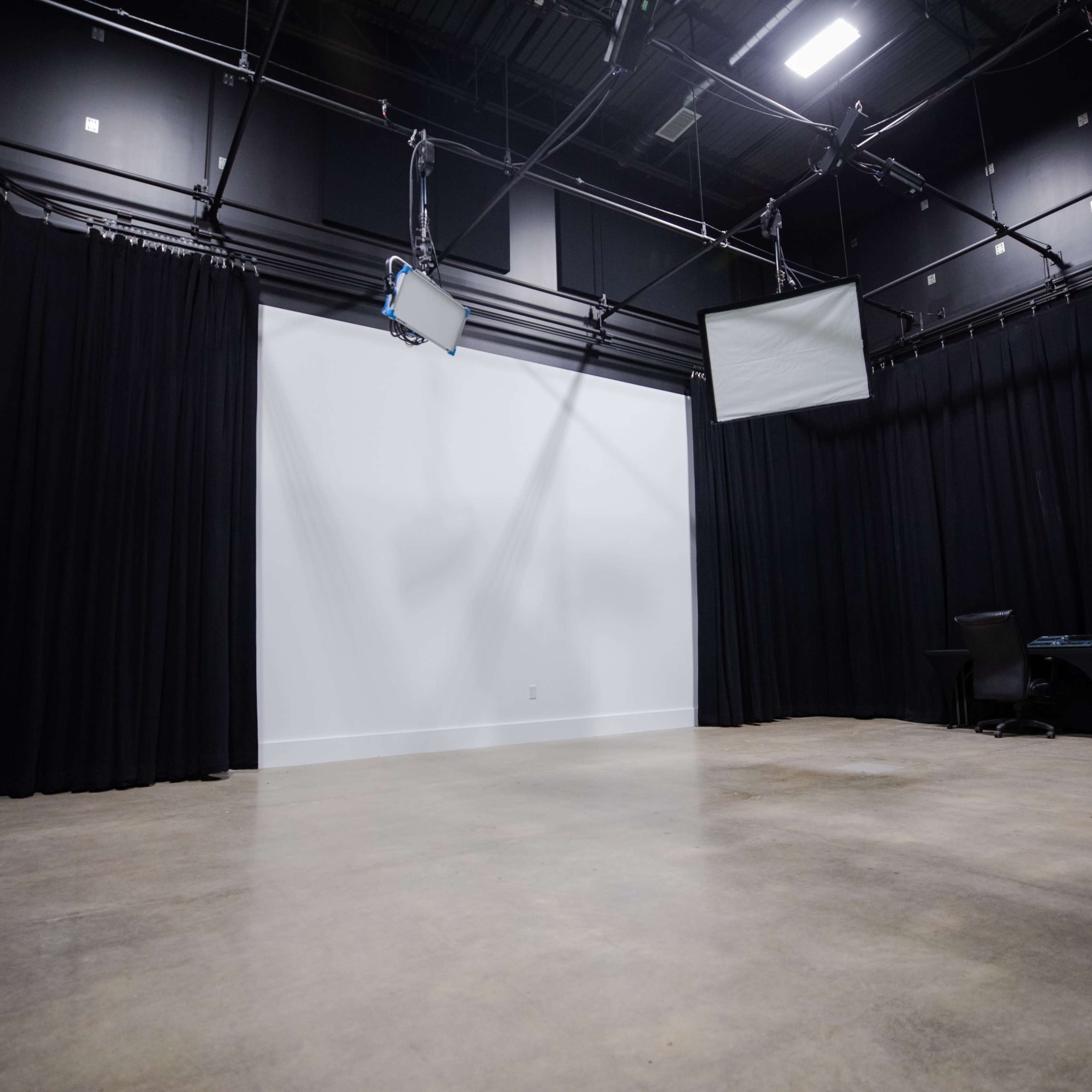 A bare studio space with black curtains, a large white wall, and two screens mounted on the ceiling.