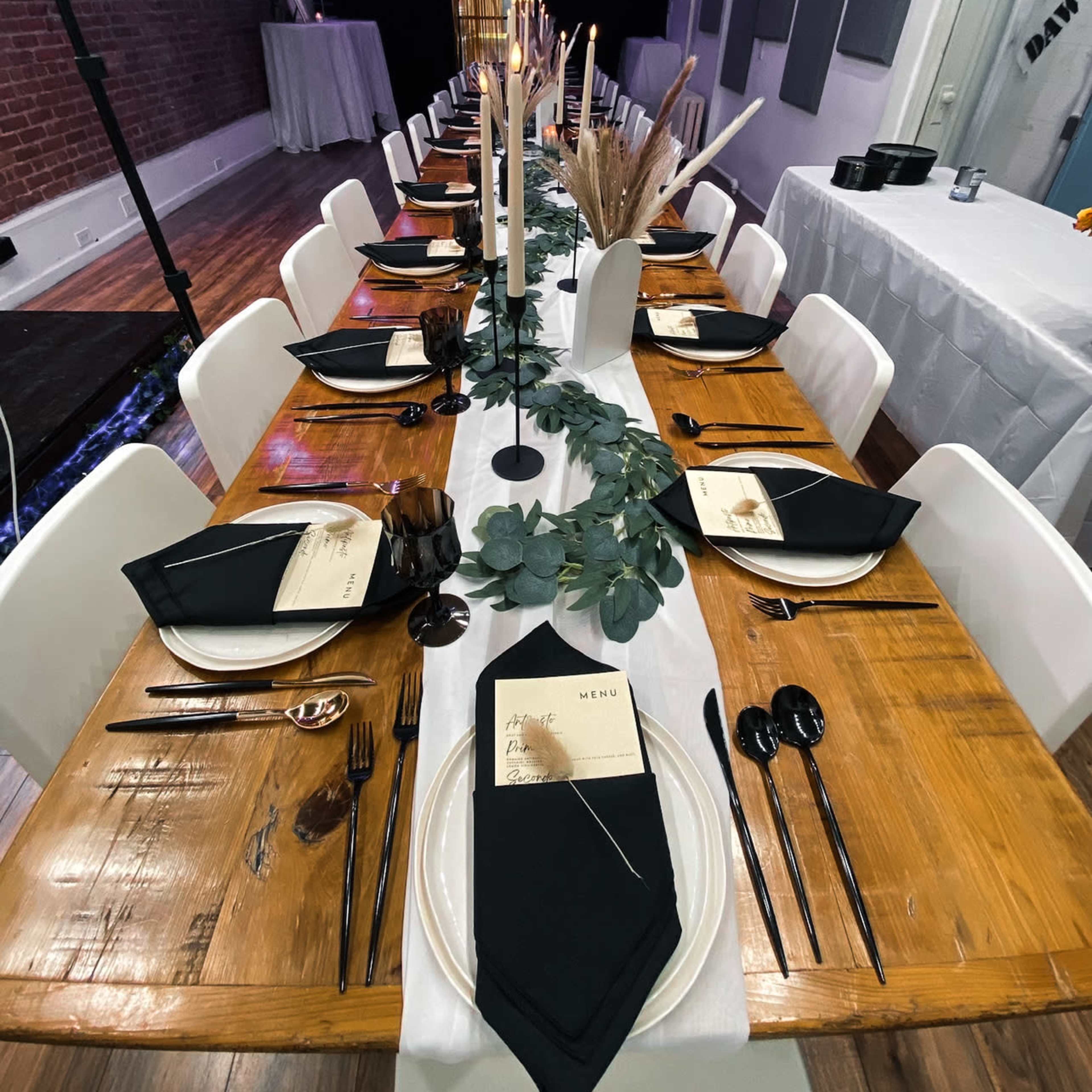 A long dining table is elegantly set with black napkins, plates, and utensils, featuring candles and greenery as centerpieces in a well-lit room.