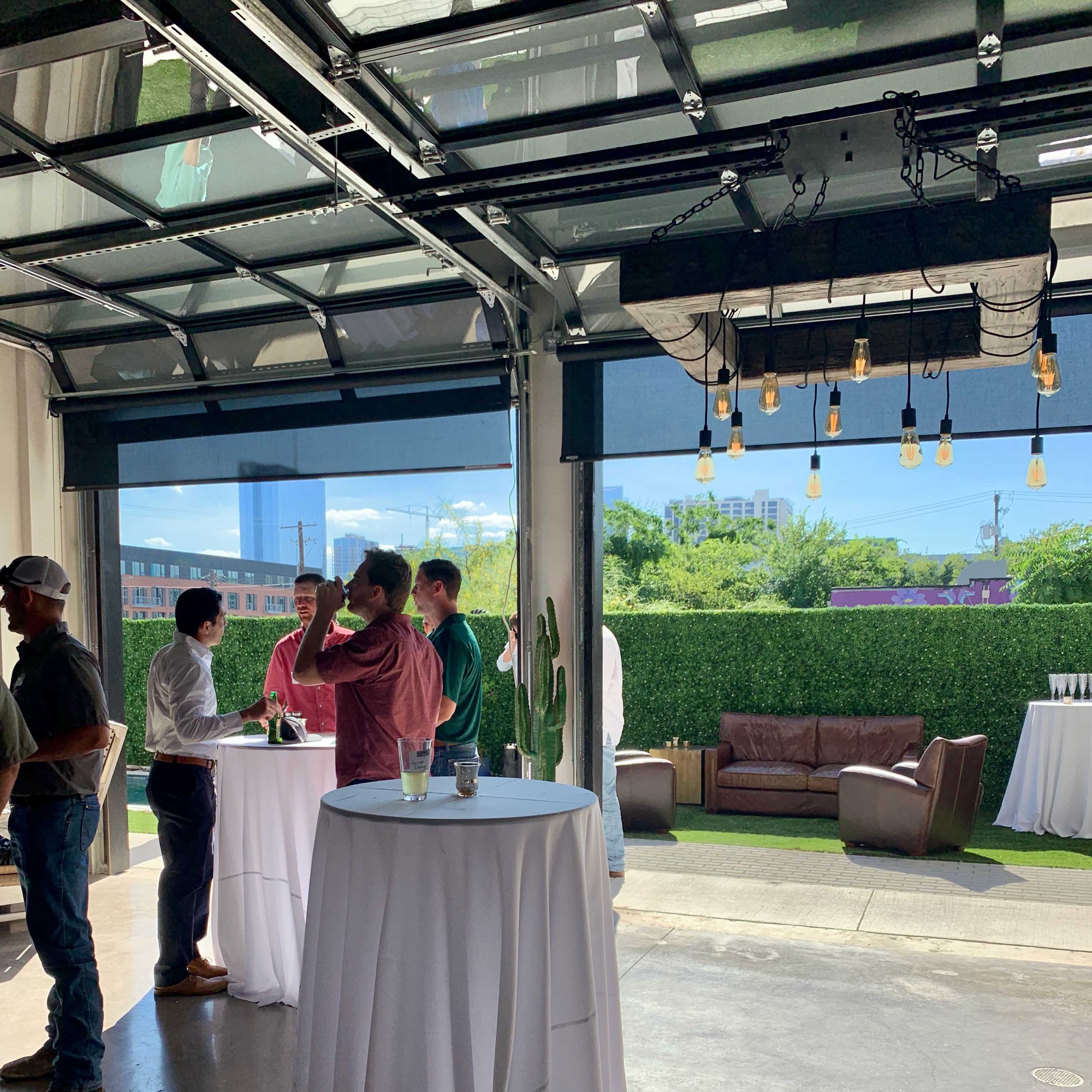 A group of people are socializing at high-top tables in a brightly lit indoor-outdoor event space with greenery in the background.