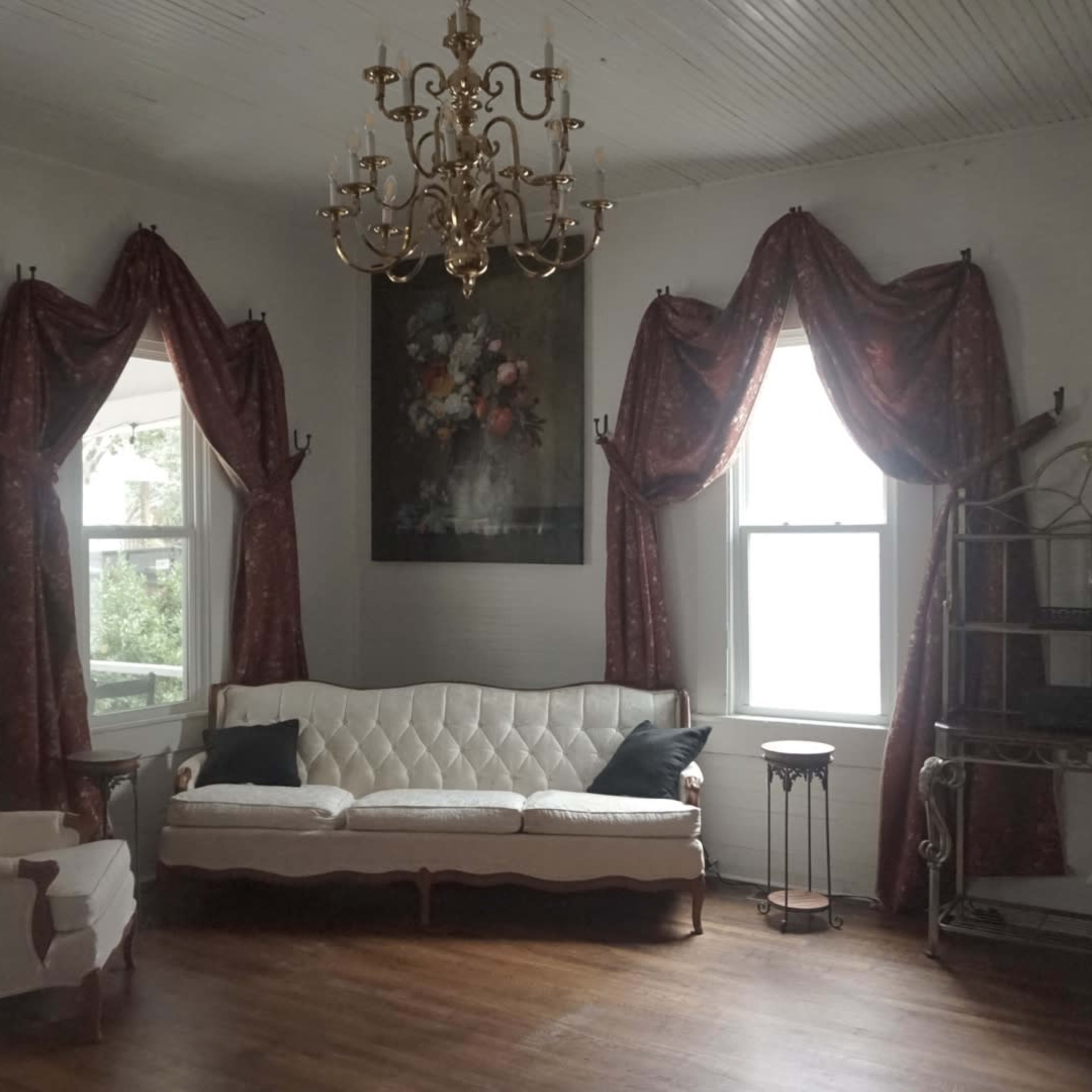 A cozy living room with a white upholstered sofa, two decorative windows with curtains, and a chandelier hanging from the ceiling.