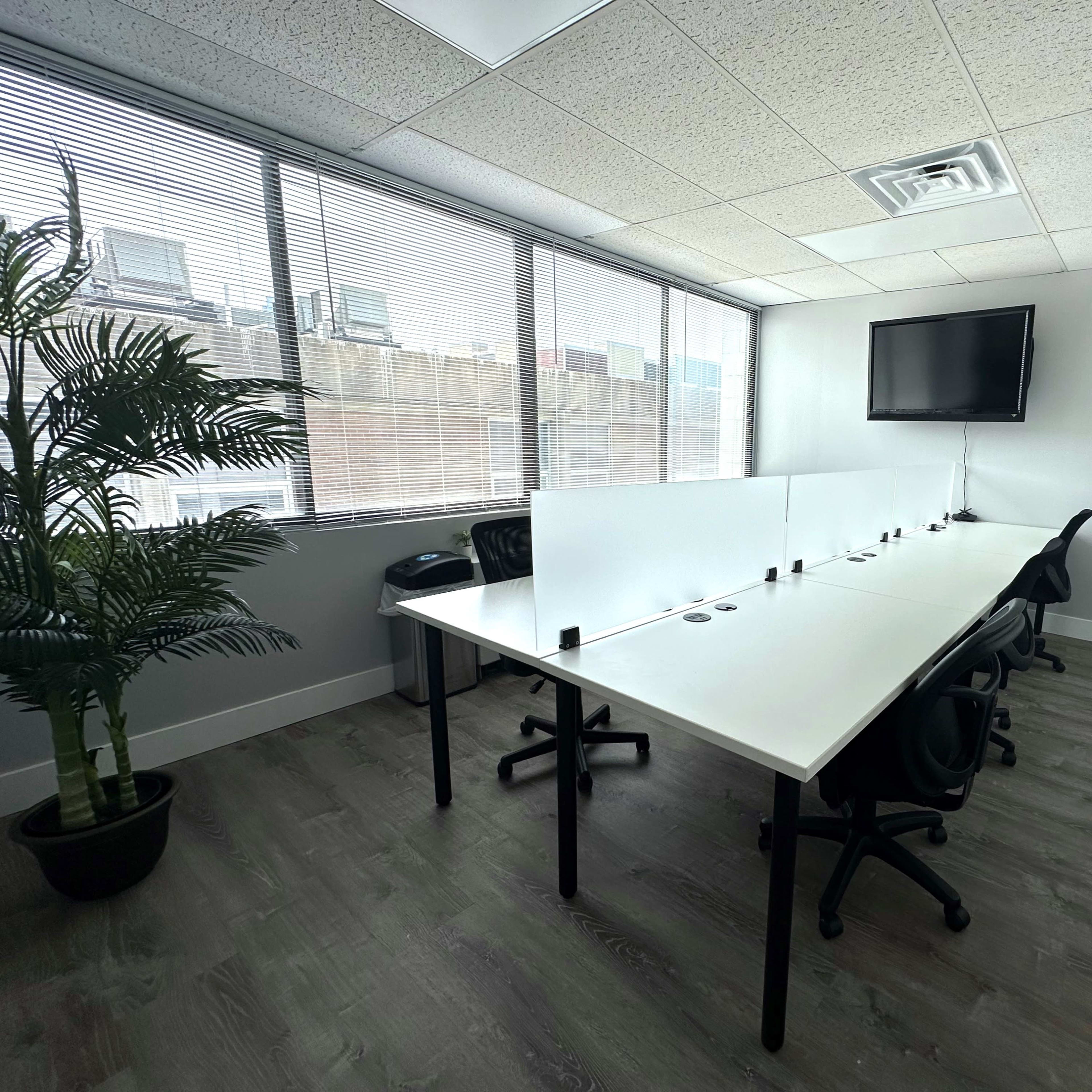 A modern conference room features a long white table with several black chairs and a wall-mounted television, illuminated by natural light from large windows.