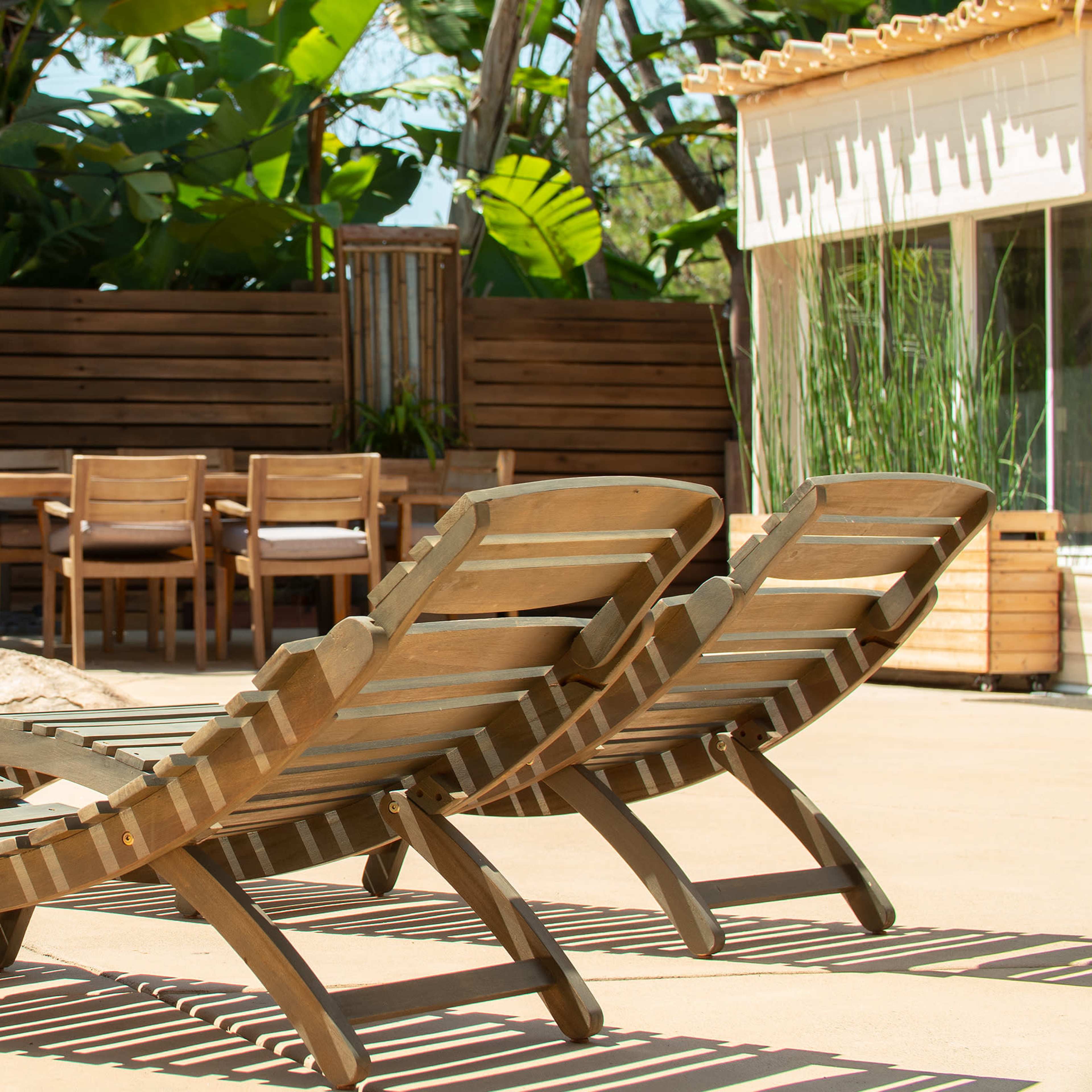 Two wooden lounge chairs positioned near a patio area with a dining table and chairs surrounded by lush greenery.