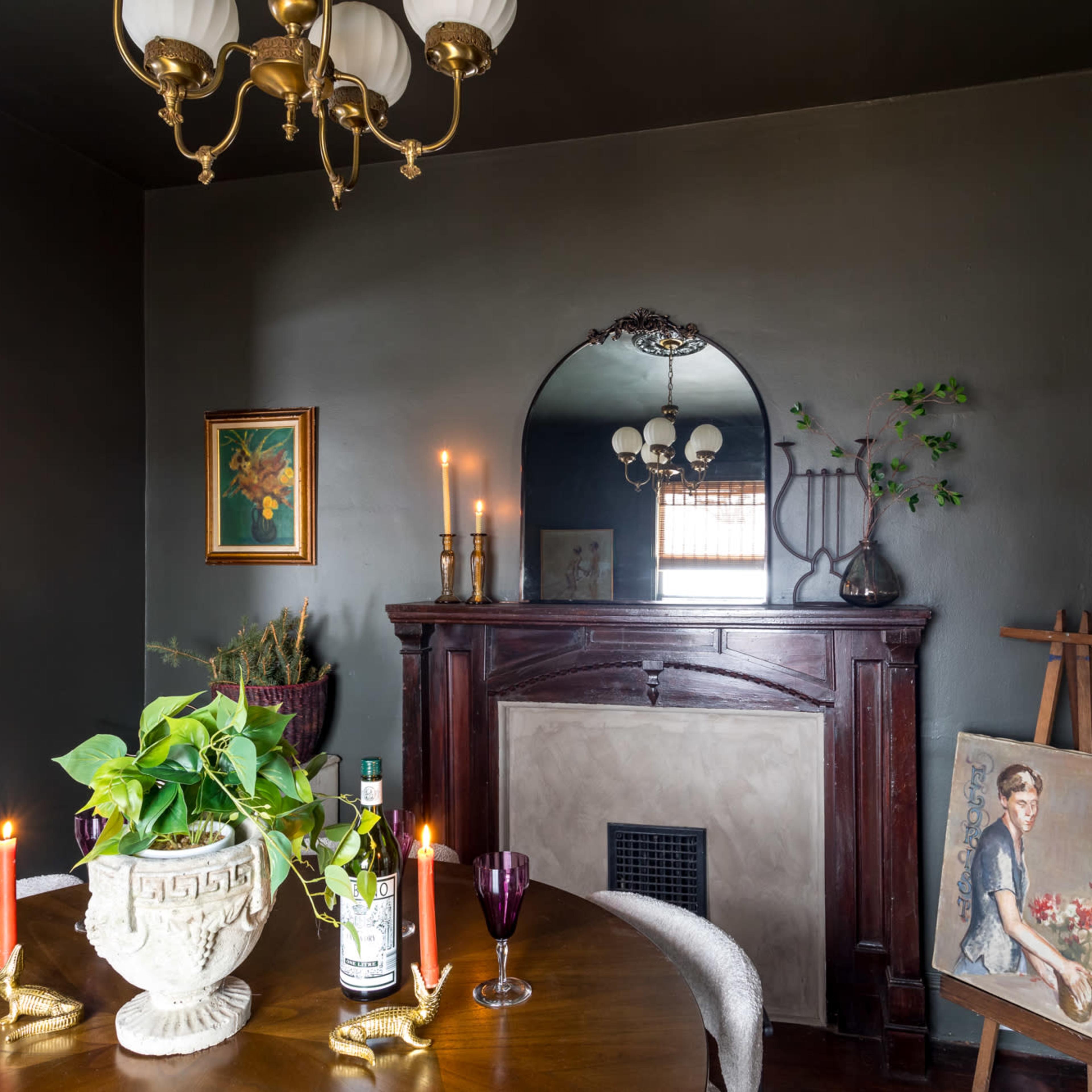 A dining area with a wooden table, a decorative fireplace, a framed painting, and a vase of greenery, illuminated by candles.
