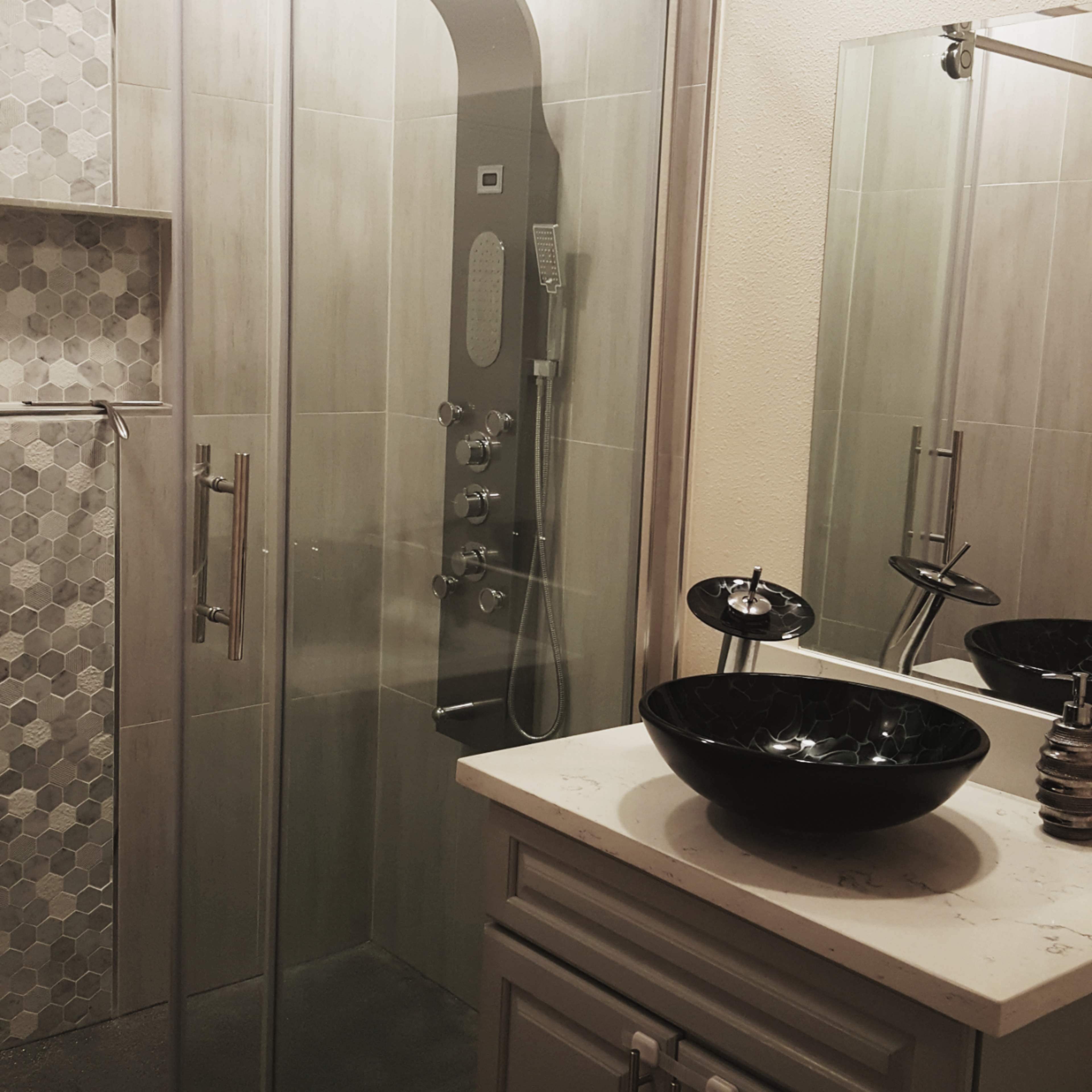 A modern bathroom featuring a glass shower enclosure with a tiled wall and a sink with a black bowl-shaped basin beside it.