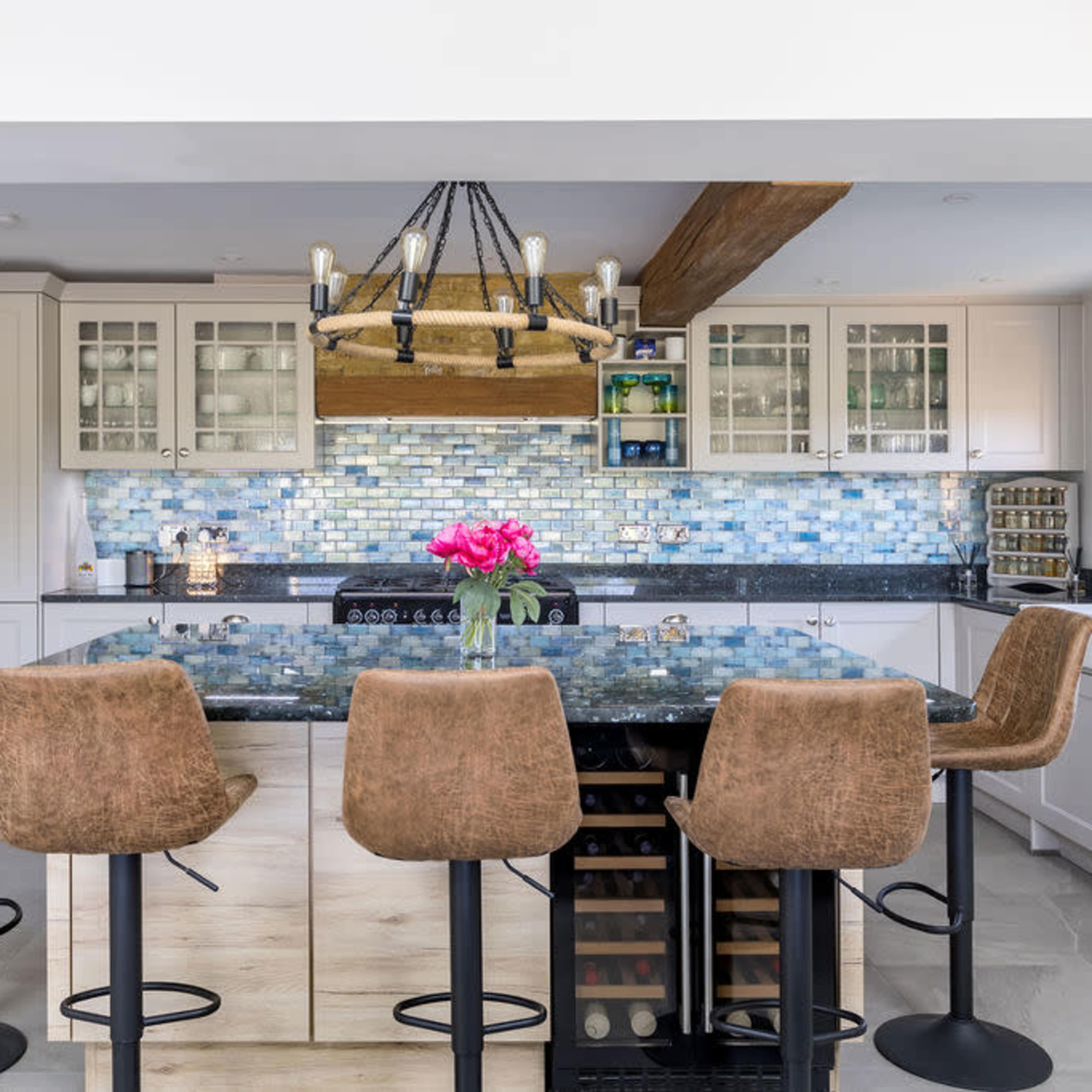 A modern kitchen features a large island with four brown bar stools, a blue mosaic tile backsplash, and a wooden beam overhead with a chandelier.
