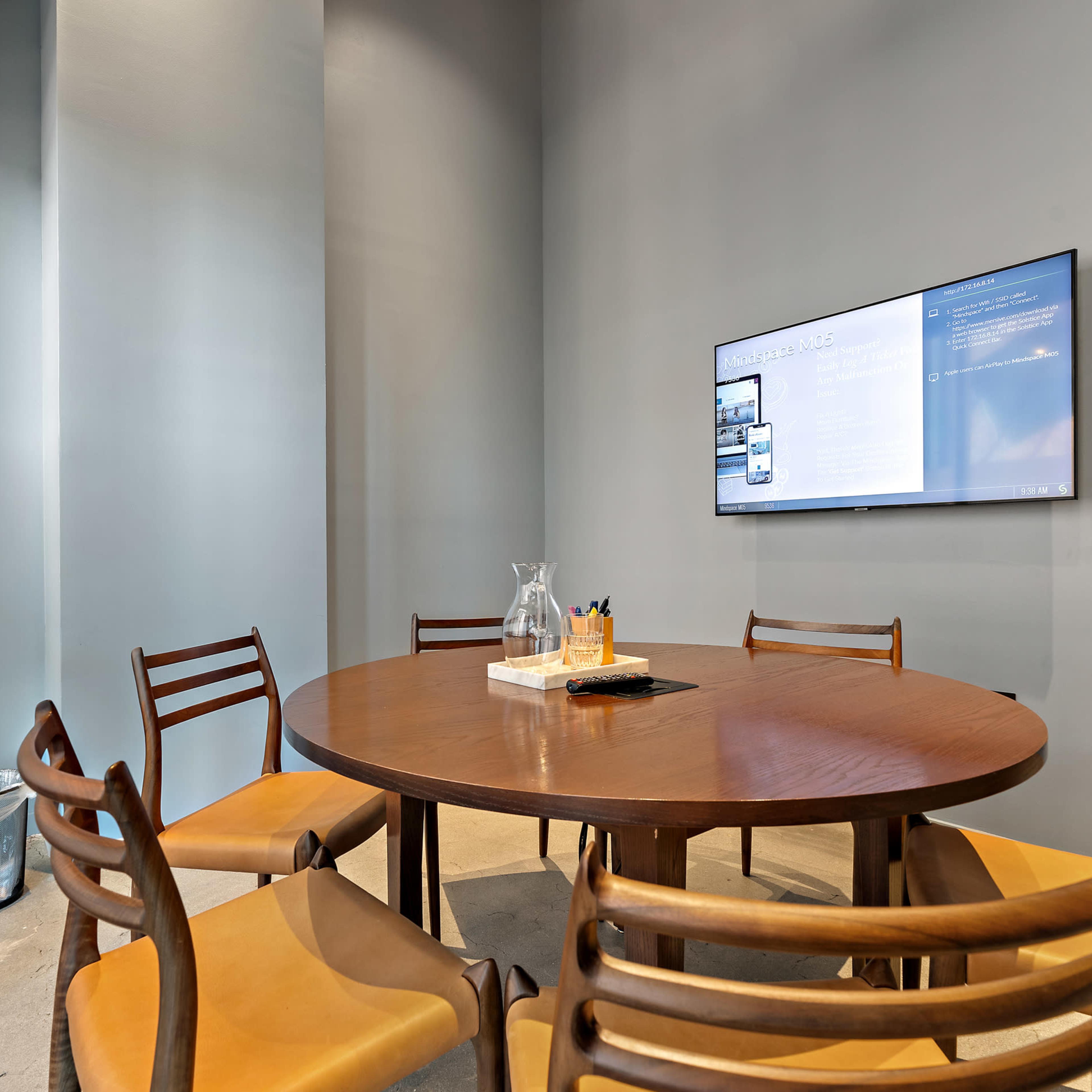 A round wooden table surrounded by six chairs is positioned in a meeting room with a wall-mounted screen displaying information.