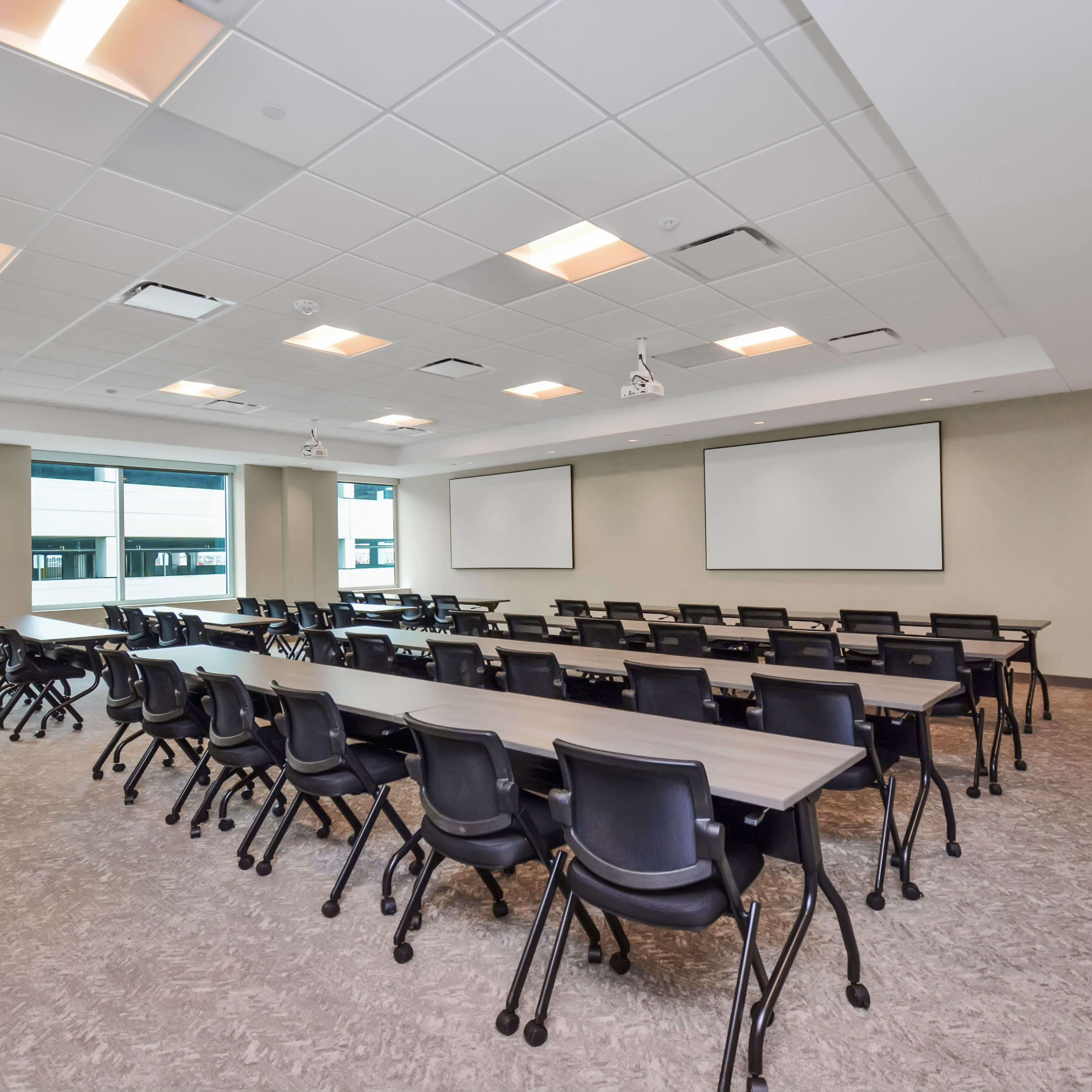 A modern conference room features several rows of black chairs facing two blank projection screens against a light-colored wall.